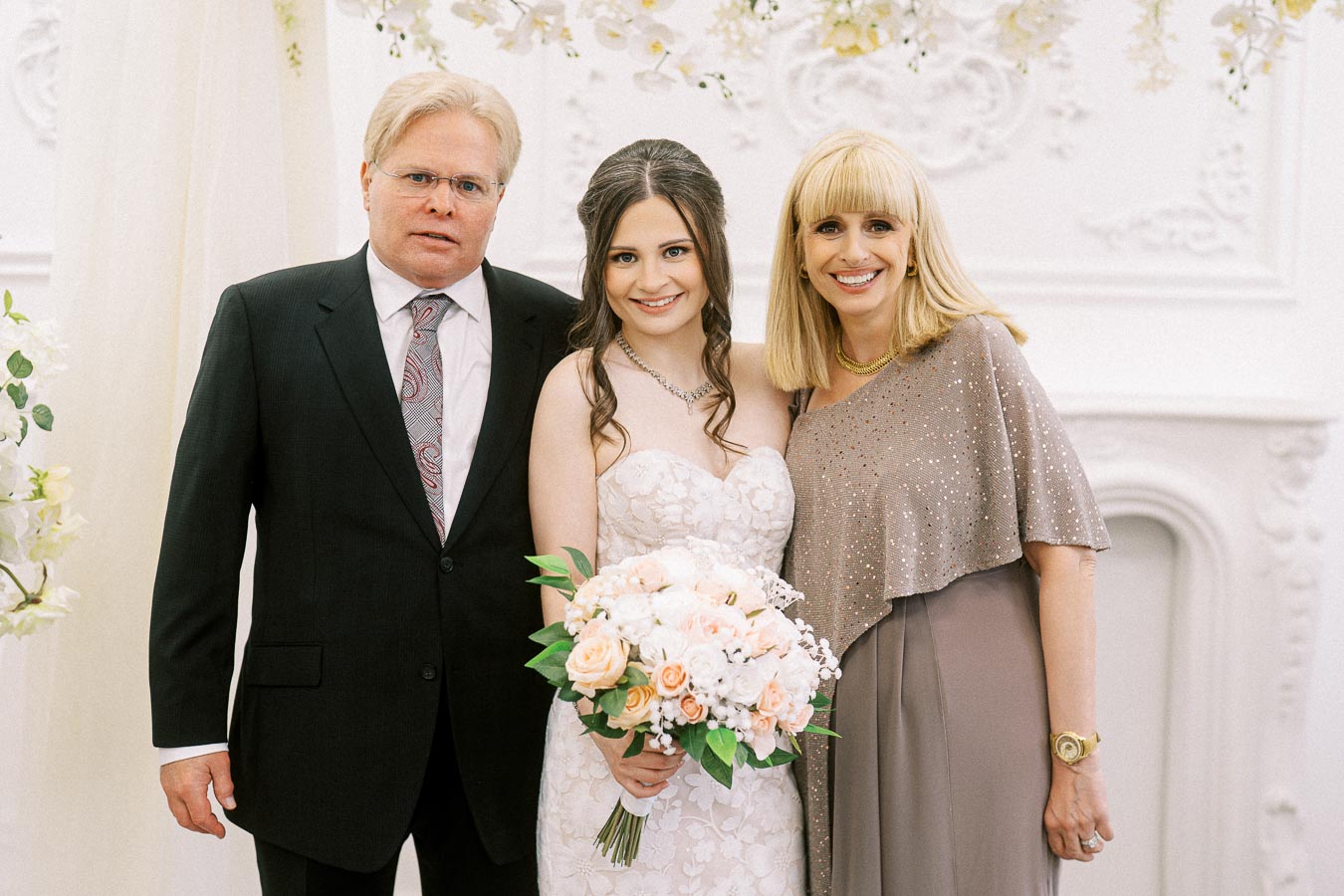 A bride in a white lace gown holding a bouquet of pink and white roses stands between two people, one in a black suit and