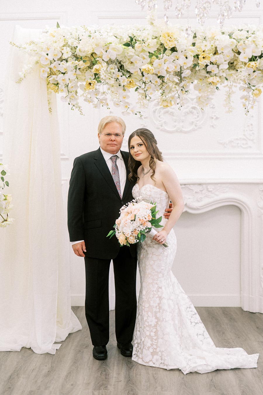 A bride in a lace wedding gown holds a bouquet standing next to a man in a black suit under a floral arch at an indoor