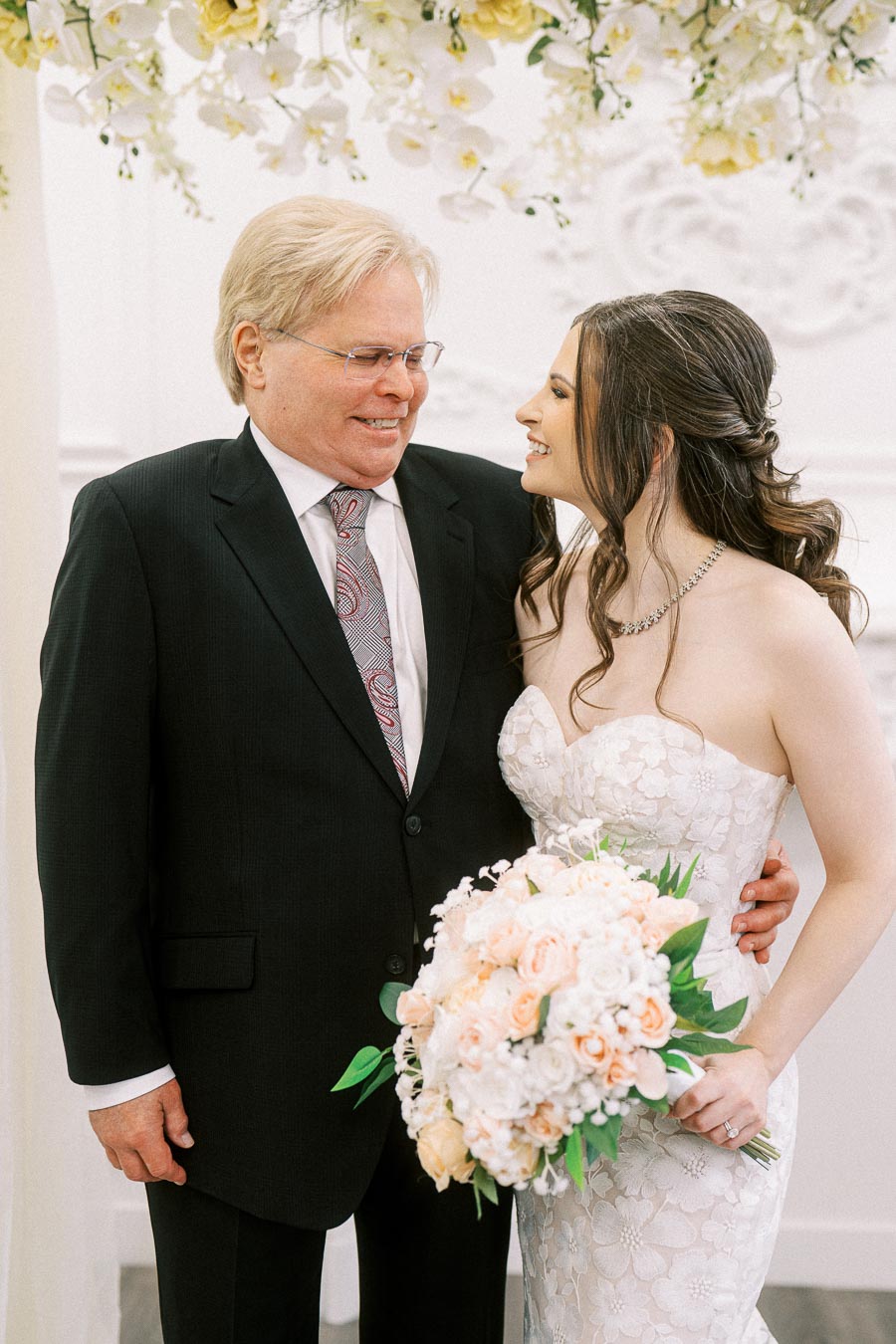 Father and bride smiling at each other during a wedding ceremony, with the bride holding a bouquet of white and peach