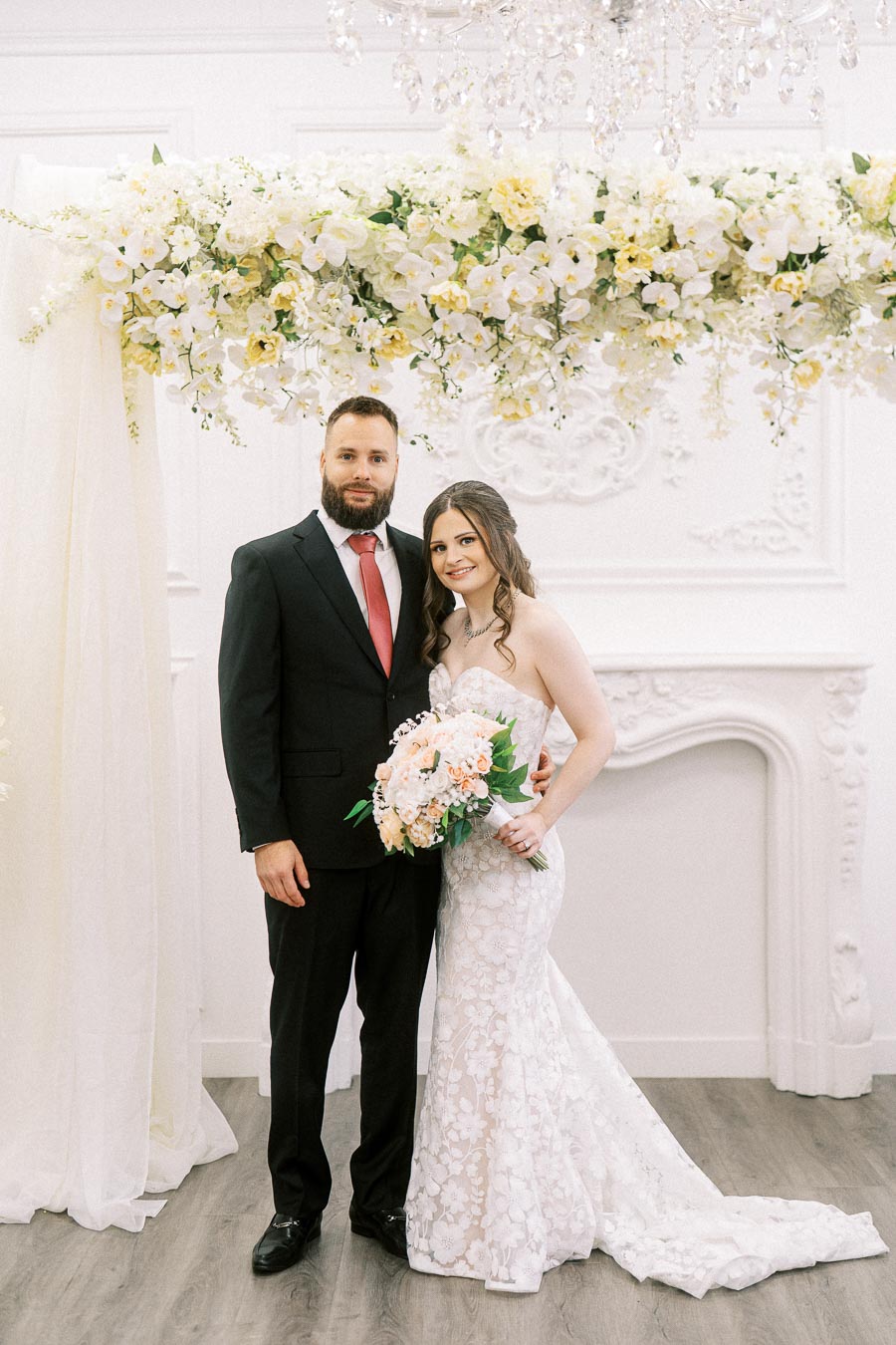A bride in a white lace wedding gown holds a bouquet of pastel flowers while standing next to a groom in a black suit and