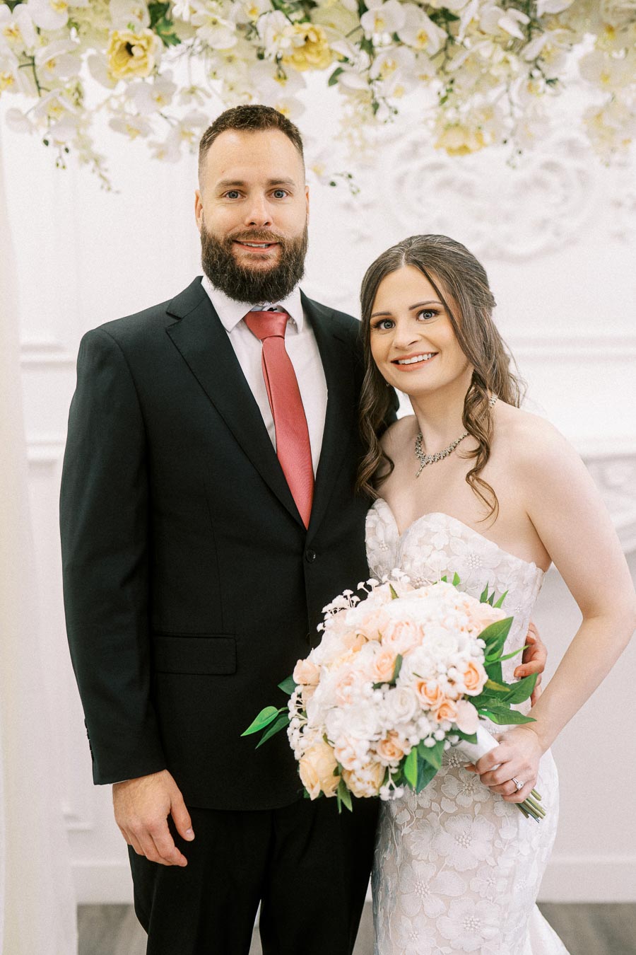 Elegant wedding portrait of a smiling bride in a lace gown holding a bouquet of pastel flowers, standing beside the groom in