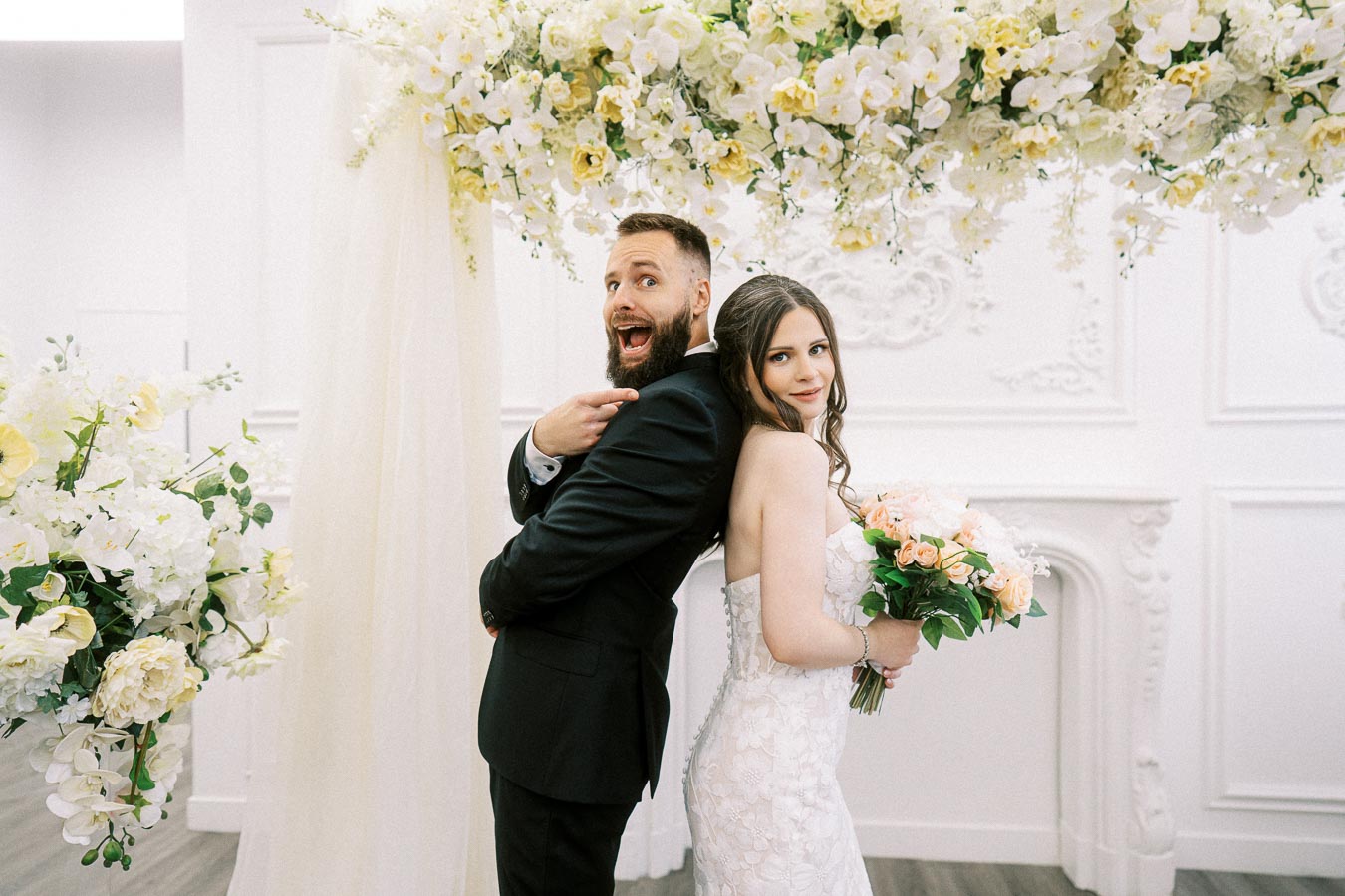 A joyful bride and groom pose back-to-back under a floral arch on their wedding day. The bride holds a bouquet of roses,