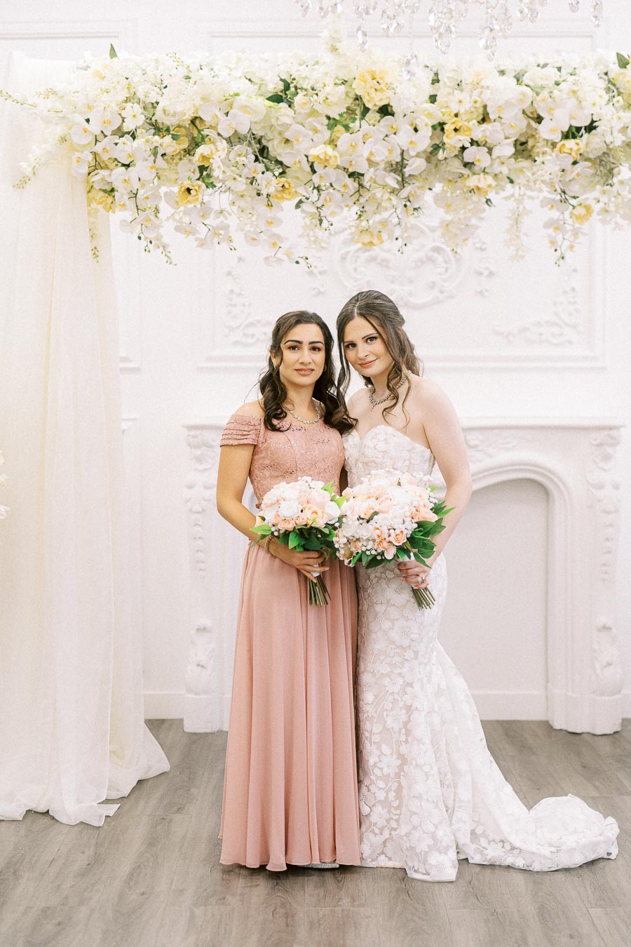 Bridesmaid and bride in elegant dresses holding bouquets under a floral arch at a wedding ceremony.