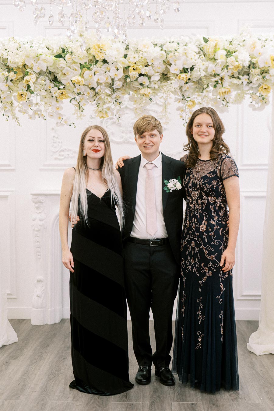 A group of three people dressed in formal attire posing in front of an elegant floral arrangement at an event. The two women