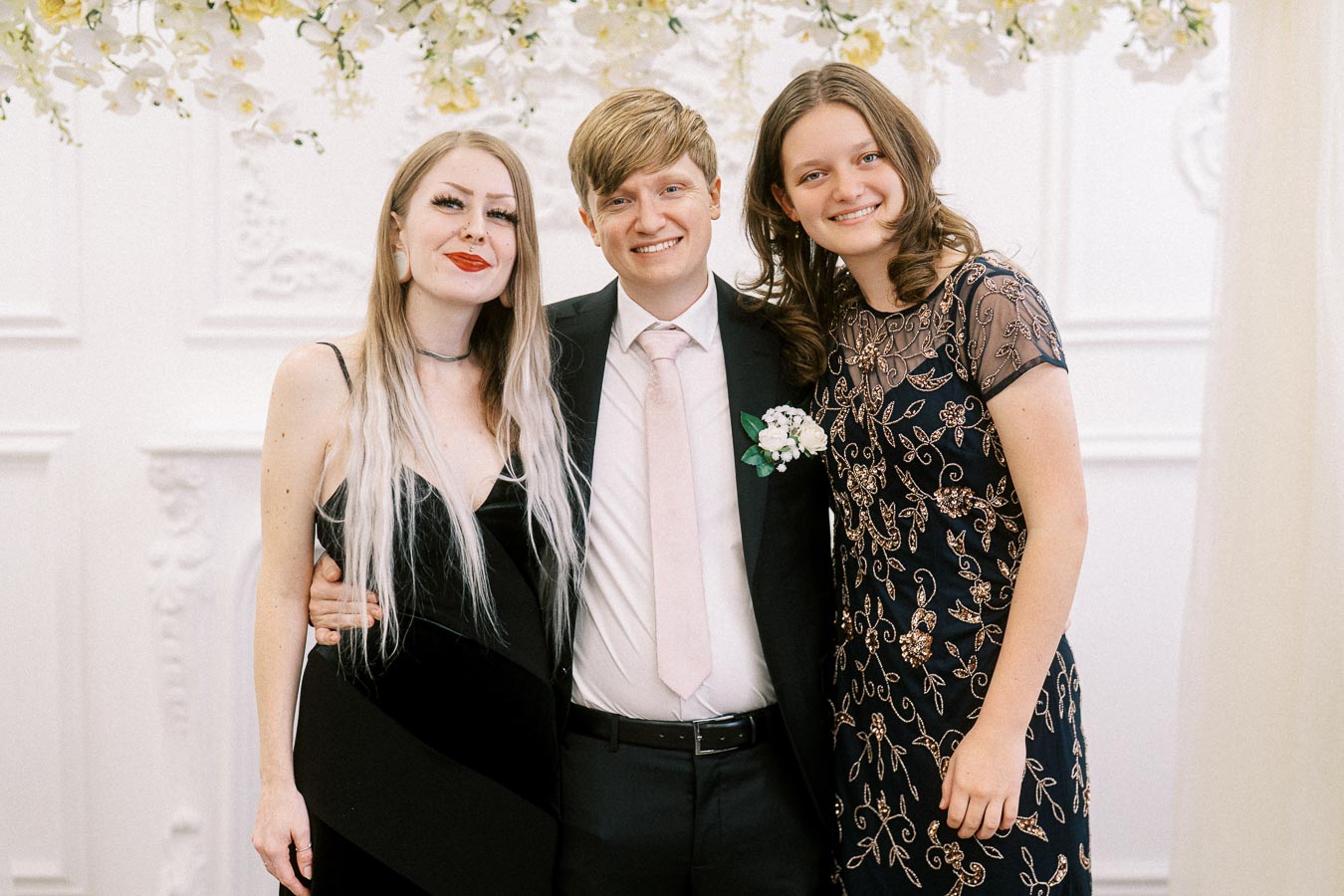 Three people posing at a formal event, featuring a man in a suit and two women in elegant dresses, smiling under a floral