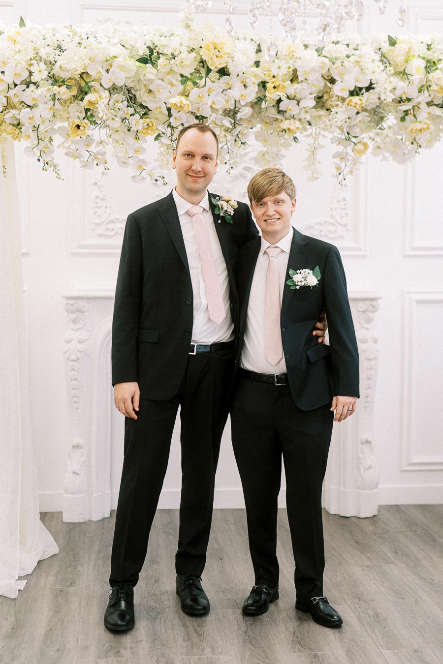 Two men in suits standing together at a wedding, adorned with floral boutonnieres and a decorative floral arch in the