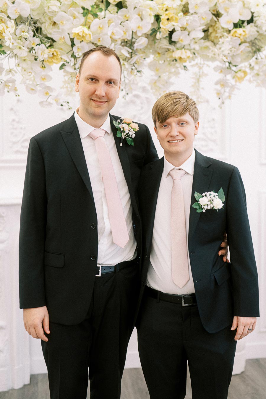 Two groomsmen wearing matching black suits and pale pink ties stand together in front of a floral backdrop at a wedding