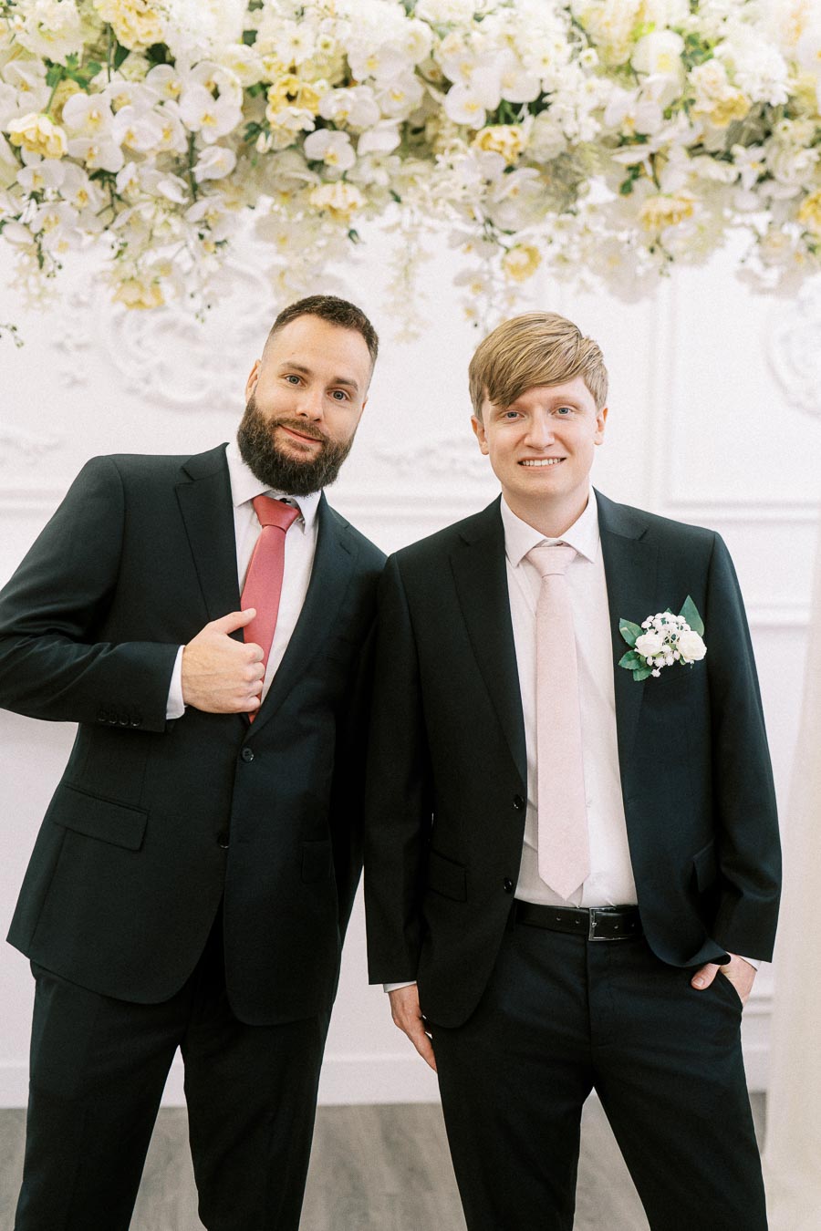 Two men in formal attire smiling at a wedding celebration, standing under a floral arrangement with white and yellow flowers.
