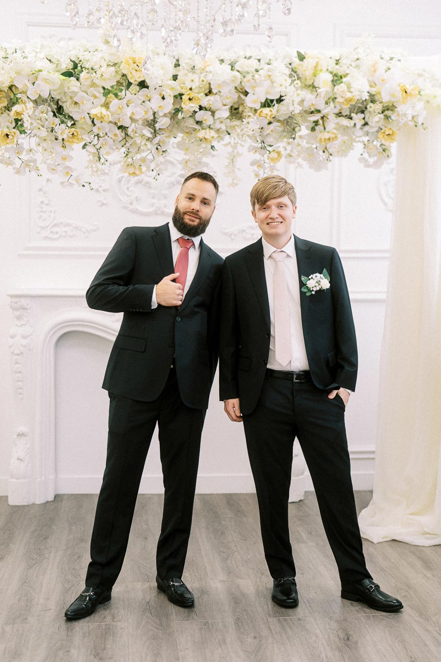 Two men in formal black suits with pink ties pose together under a floral wedding arch in an elegantly decorated room.