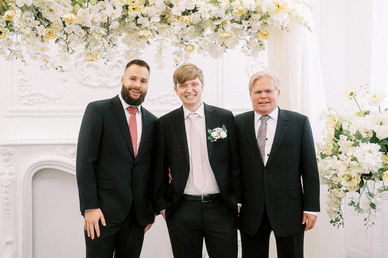 Three men in suits standing together at a wedding ceremony, smiling for a group photo beneath an elegant floral arch with
