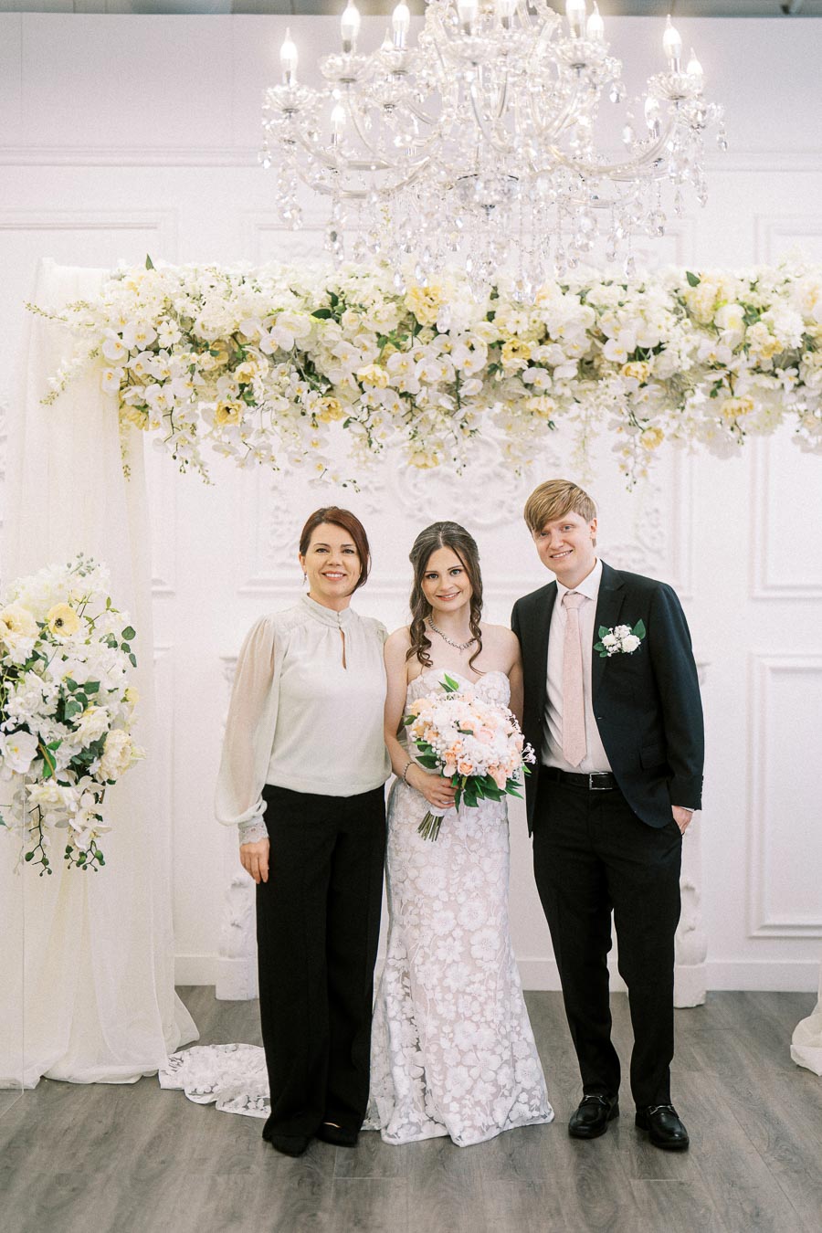 A bride and groom standing with a woman in a wedding venue, featuring elegant floral arrangements and a crystal chandelier,