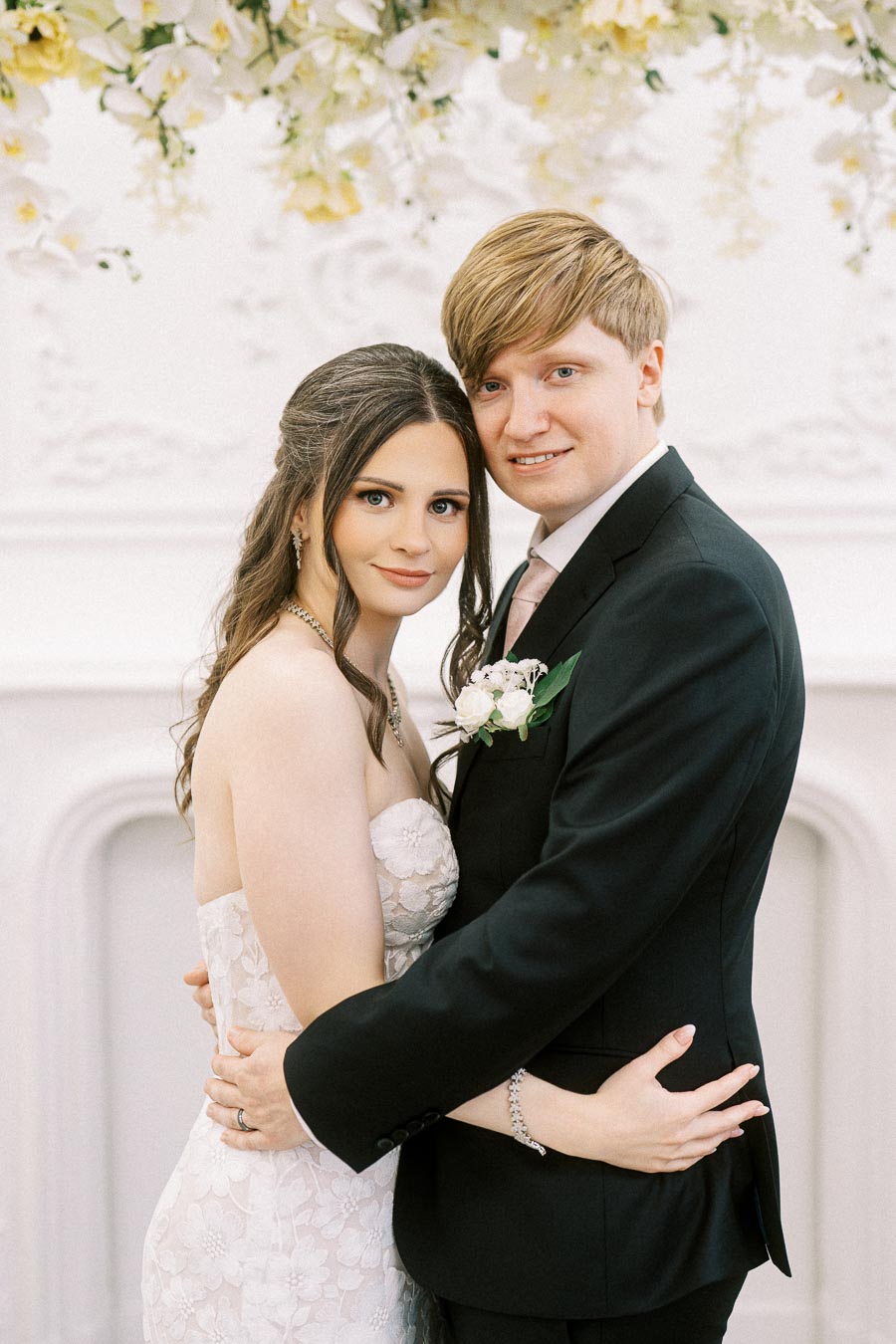 A newlywed couple embracing on their wedding day, with the bride in a lace strapless gown and the groom in a black suit,