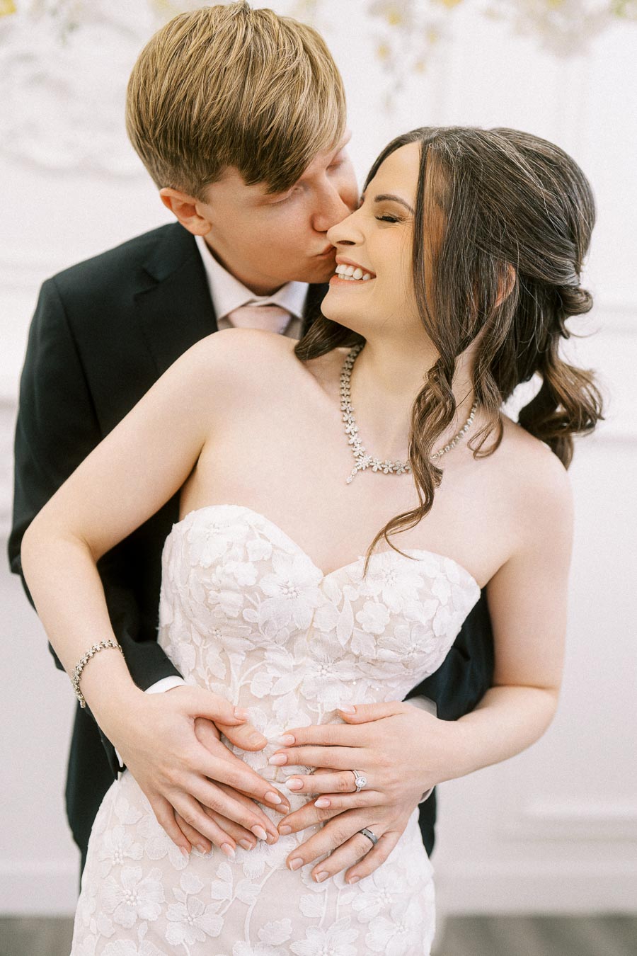 Happy couple embracing on their wedding day, with the groom in a black suit kissing the smiling bride in a white floral lace