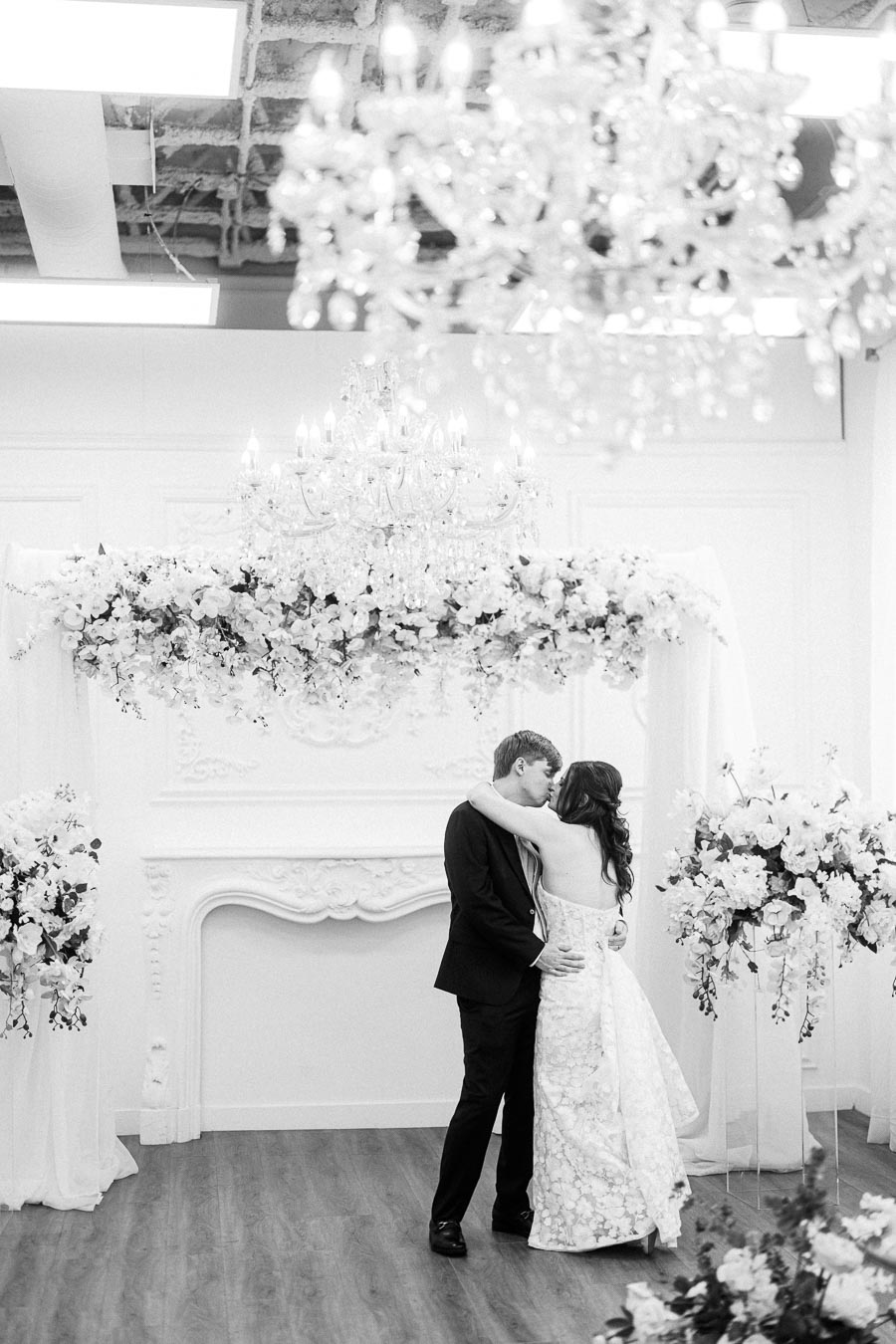A romantic black and white wedding photo capturing a couple embracing under an elegant floral arch and chandelier in a