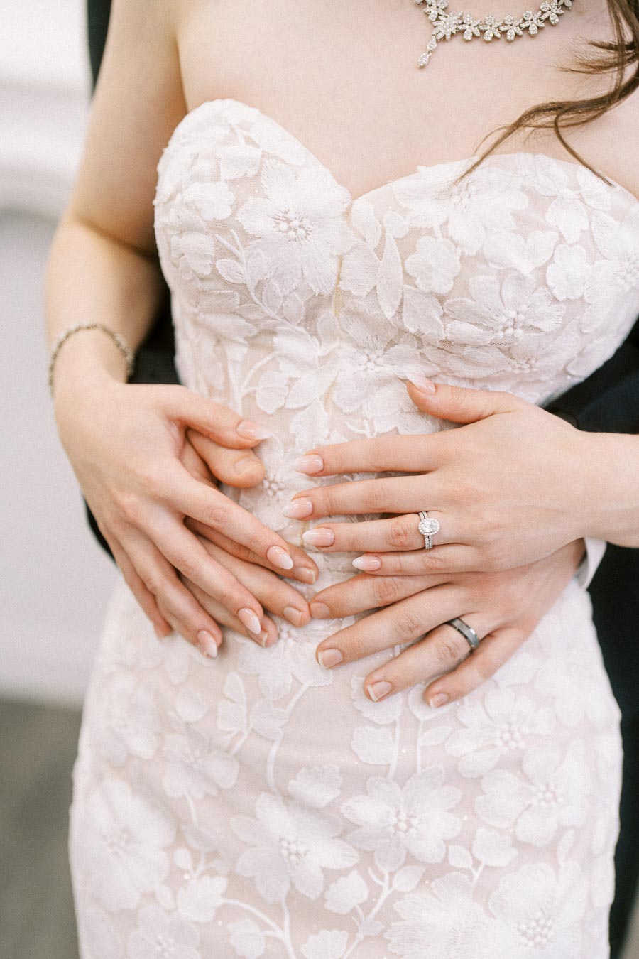 Close-up of a bride's white floral lace wedding dress, with hands intertwined displaying wedding rings, symbolizing love and