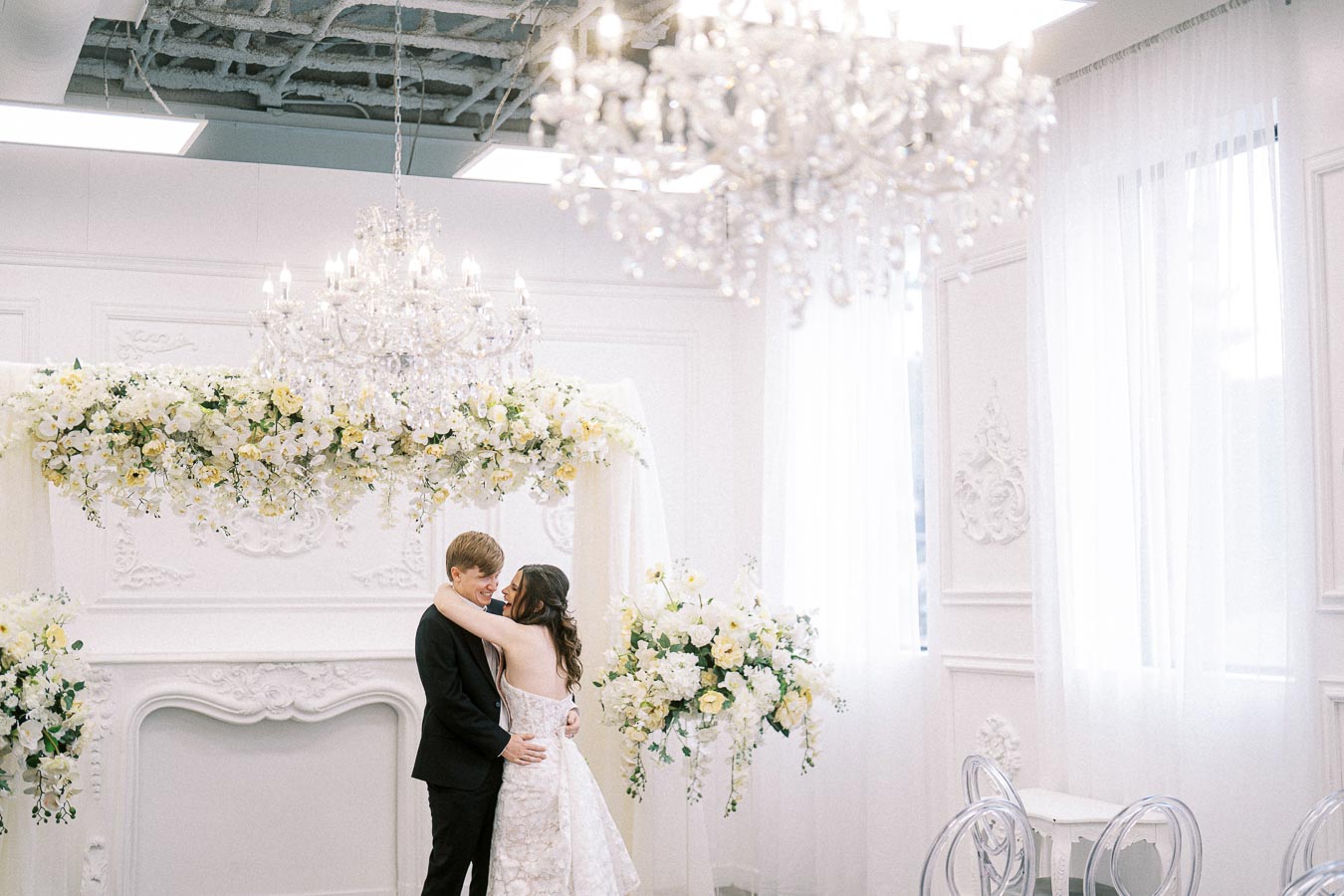 A couple embraces under an elegant chandelier in a beautifully decorated white room with floral arrangements, capturing a