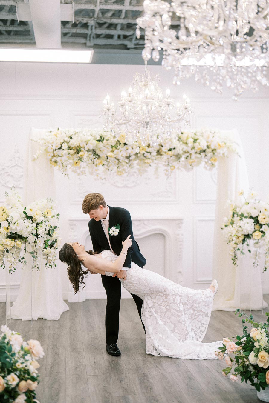 A bride and groom share a romantic dance in an elegant wedding venue adorned with chandeliers and white floral arrangements.