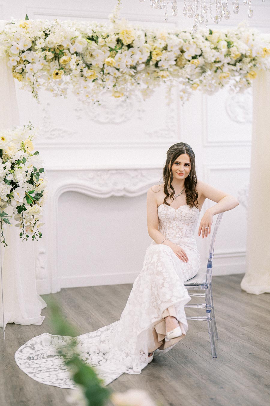 A bride in an elegant lace wedding dress sits on a transparent chair, surrounded by floral decorations with white and yellow