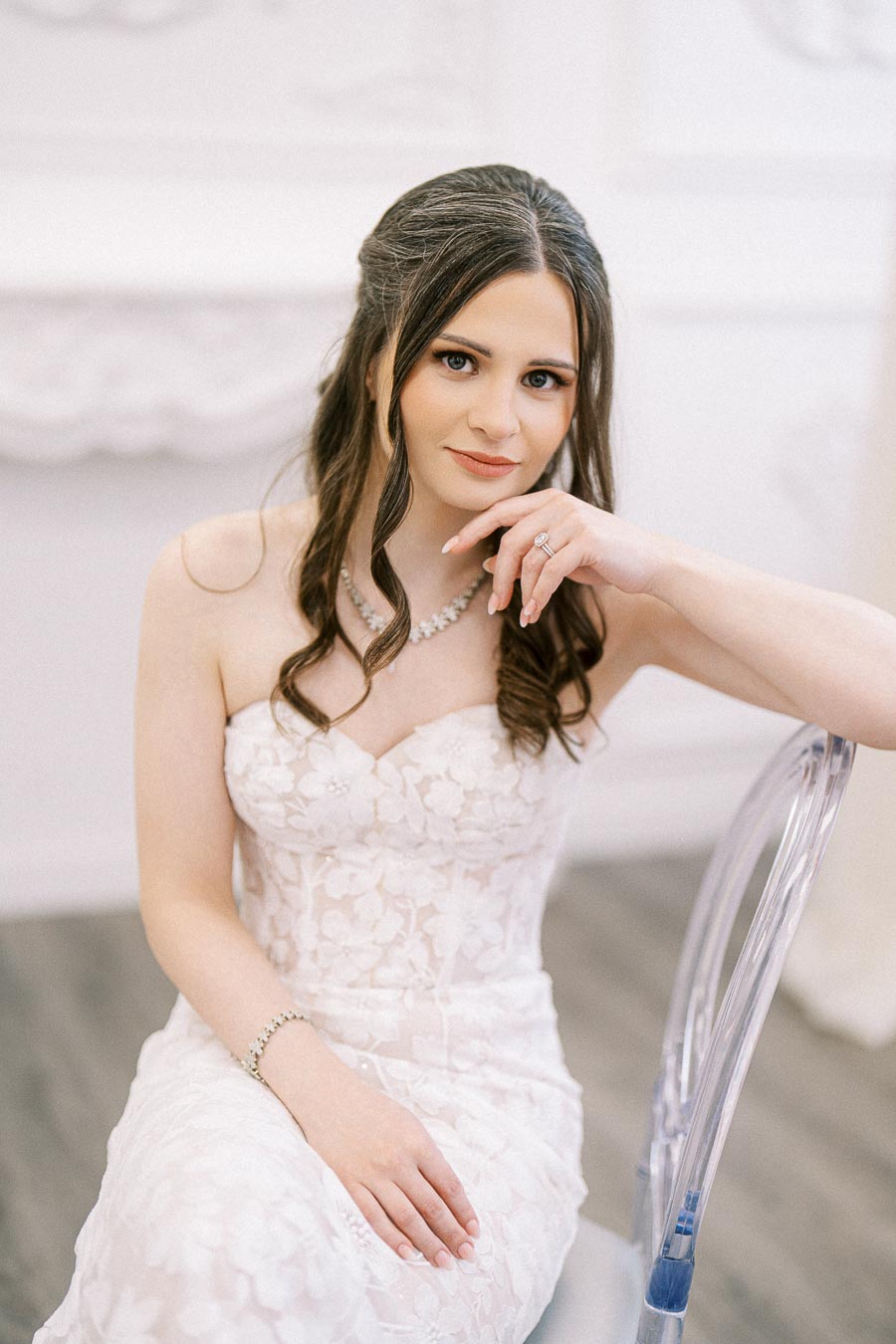 Bridal portrait of a woman in an elegant white floral lace wedding dress, seated on a clear chair, with long wavy hair and