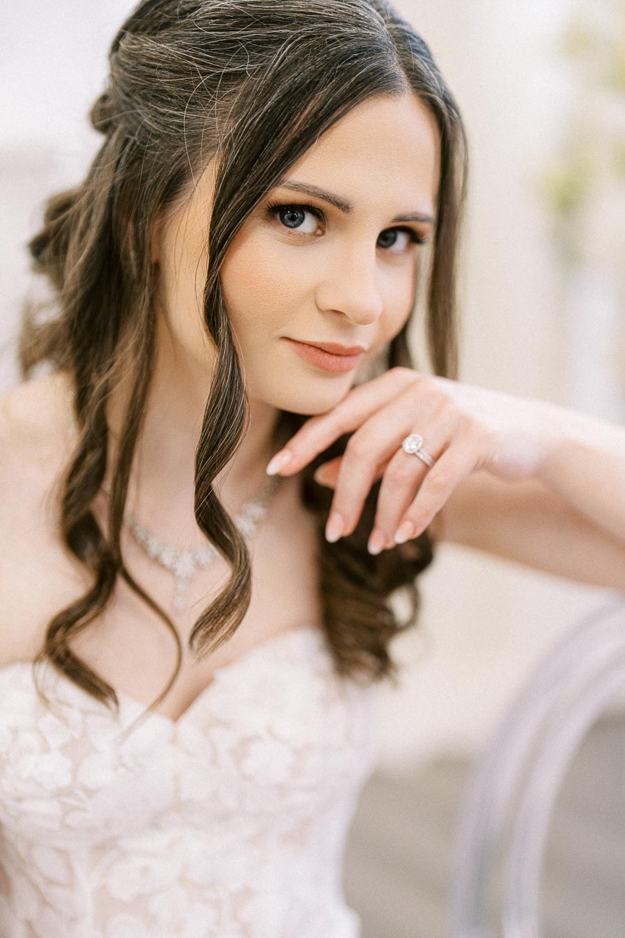A bride with long dark hair styled in waves, wearing a strapless wedding gown with floral lace details, poses gracefully,