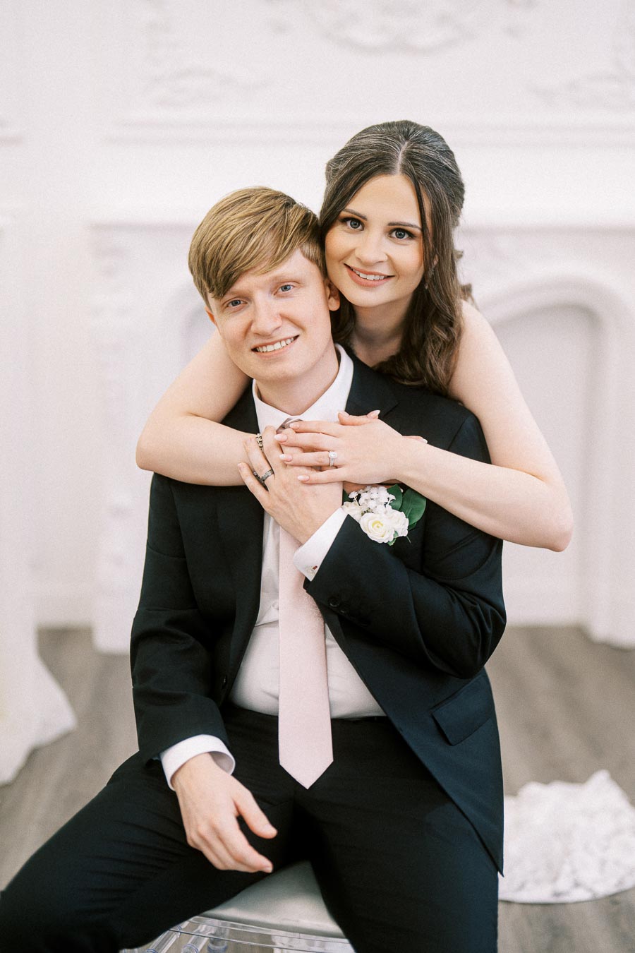 A happy couple dressed in wedding attire, with the bride embracing the groom from behind. The groom is seated wearing a
