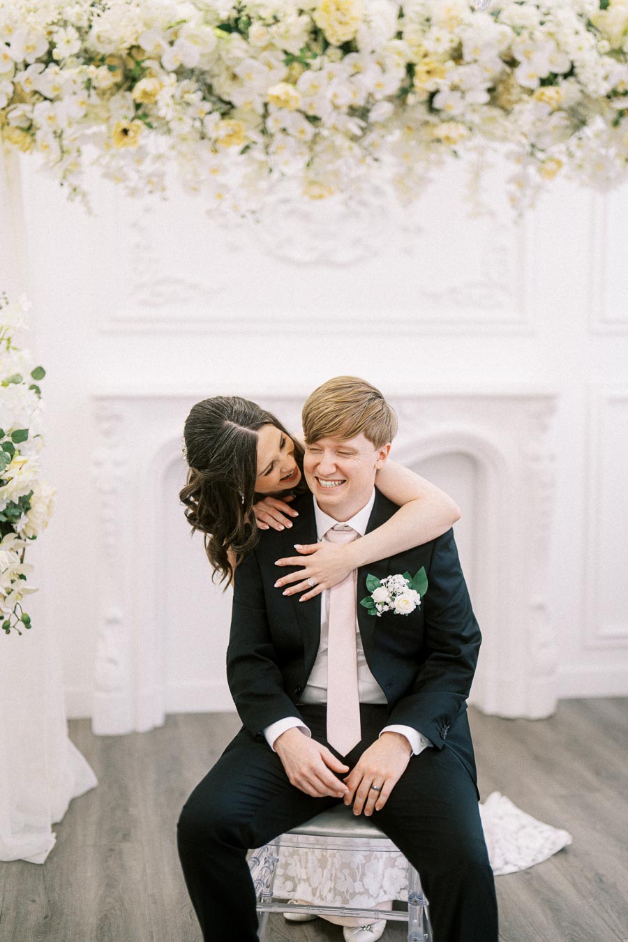 Happy couple embracing during wedding ceremony, surrounded by elegant floral decor, with groom in a black suit and pink tie,