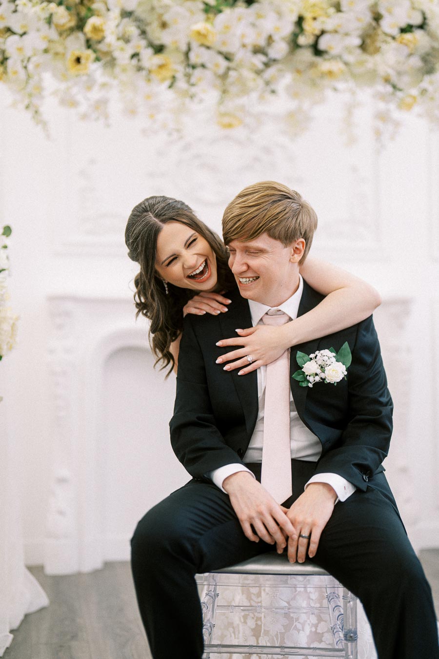 A joyful couple on their wedding day, with the bride embracing the groom from behind as they share a laugh in a beautifully