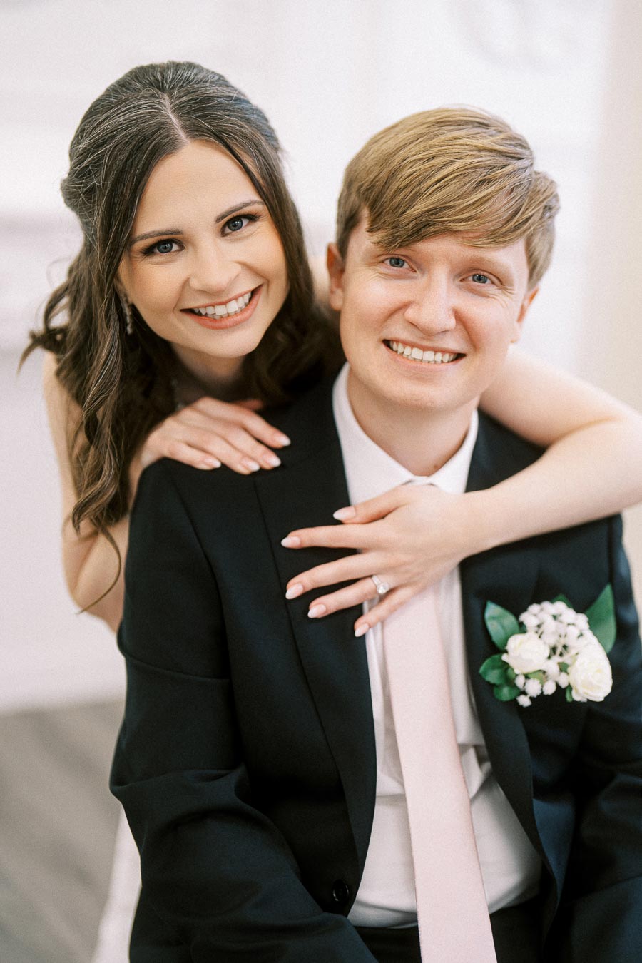 Smiling bride and groom embracing, wearing formal wedding attire with floral boutonniere.