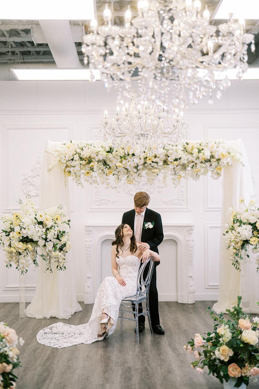 Elegant wedding photoshoot with a bride in a white lace gown seated under a chandelier, surrounded by white and yellow