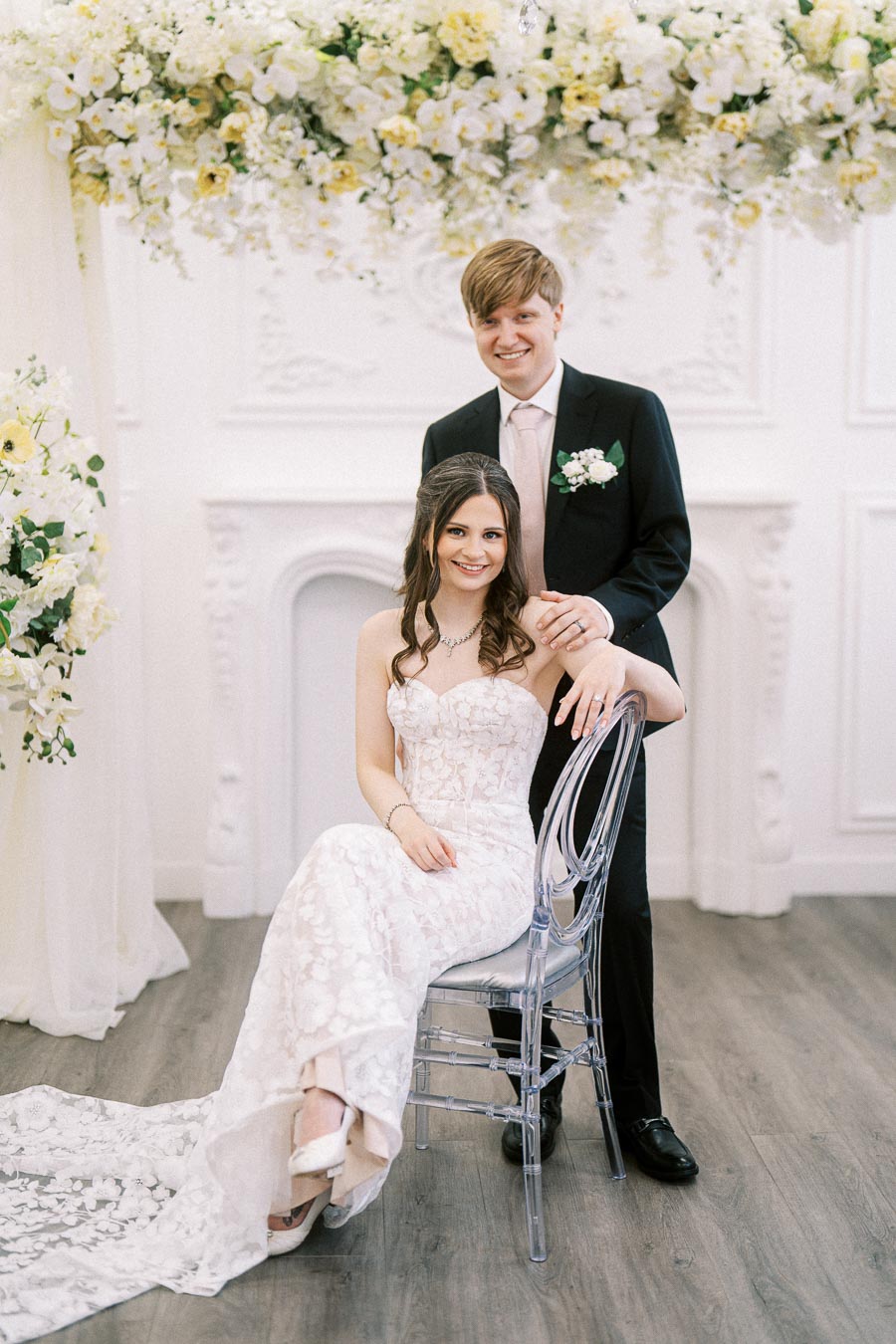 A bride in a white lace wedding dress sits on a clear chair, while the groom, wearing a black suit with a floral