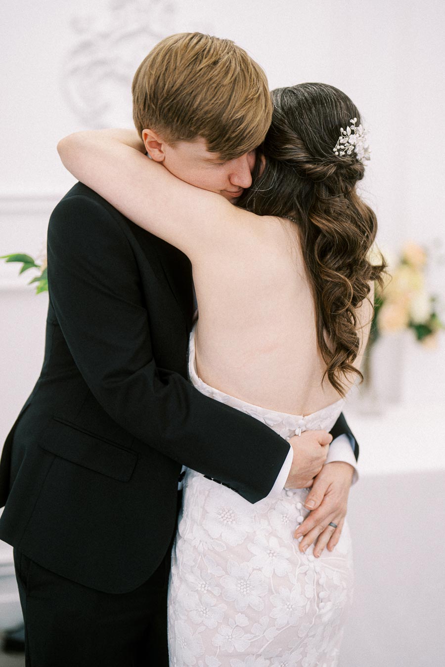 A bride and groom share a heartfelt embrace in a bright room, highlighting the bride's elegant floral lace wedding gown and
