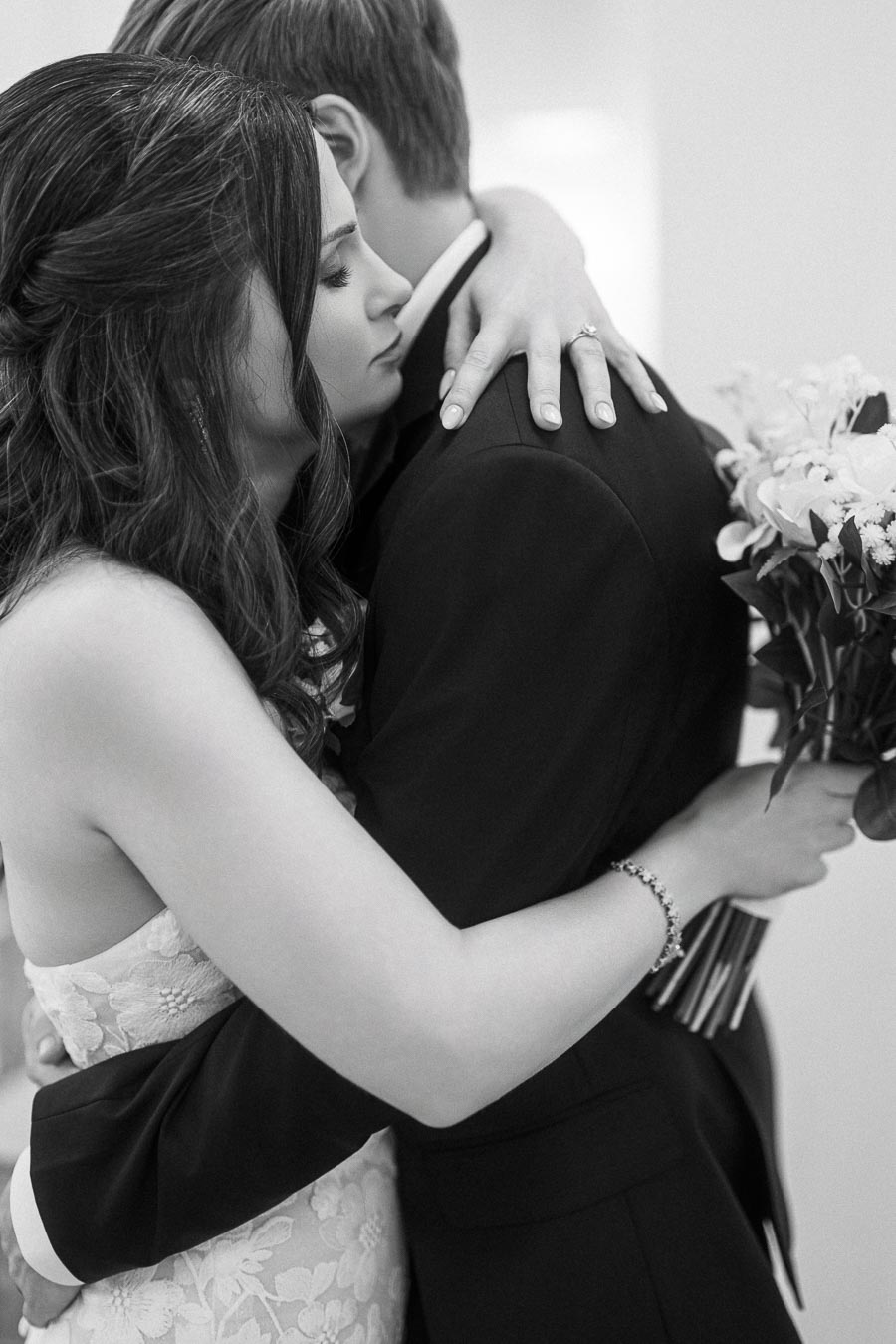 A black and white photo of a couple embracing, with the woman in a lace dress holding a bouquet of flowers, capturing an