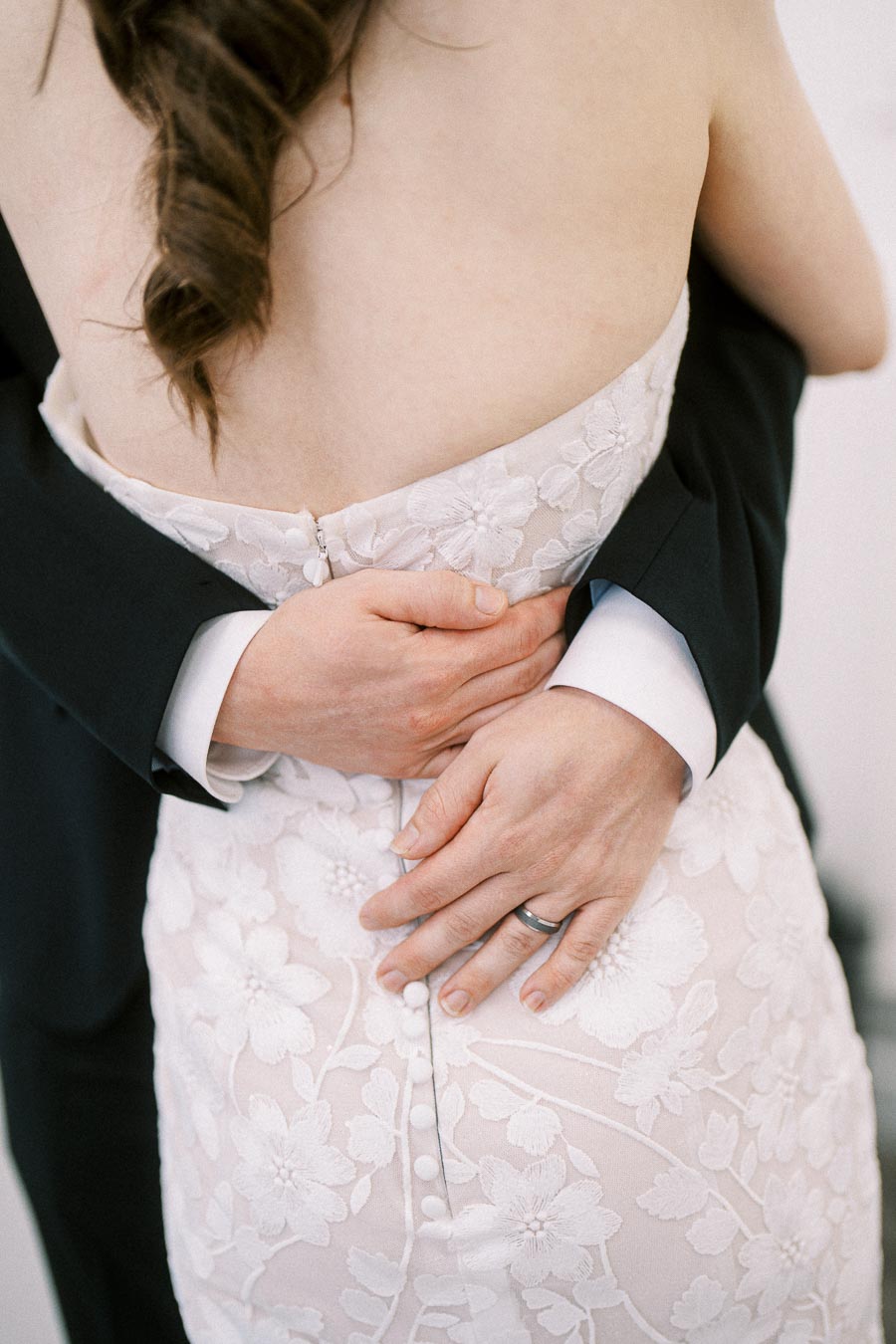 Bride and groom embrace closely, focusing on hands and floral lace wedding dress detail.