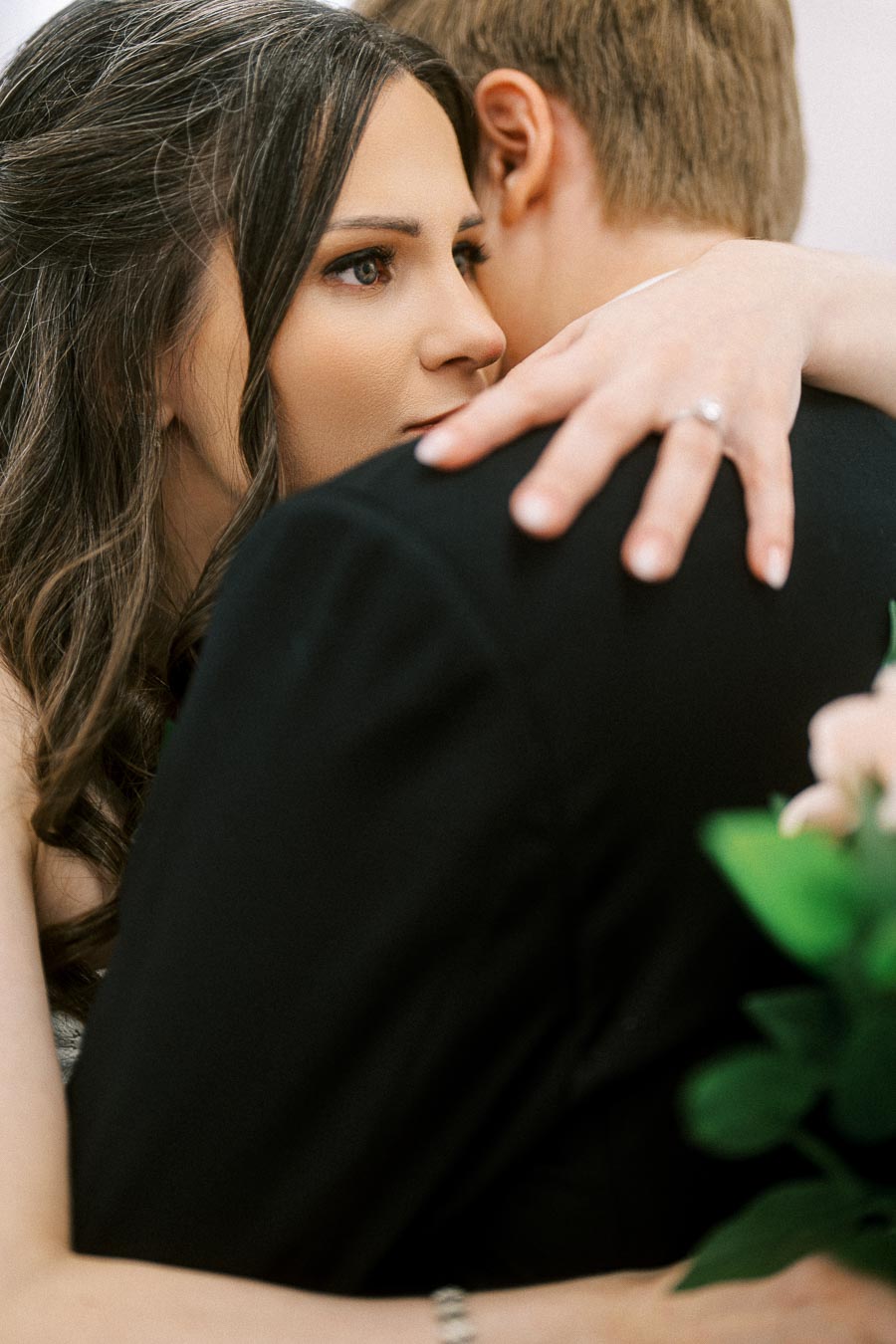 A close-up of a bride embracing her partner, focusing on her hand with an engagement ring and lush bouquet of green leaves.