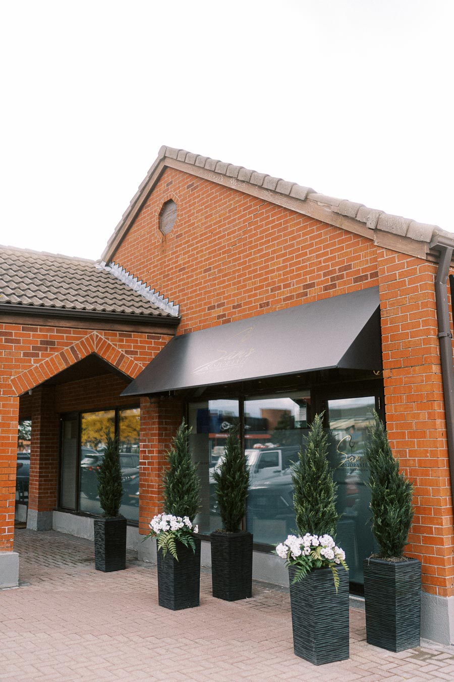 Exterior view of a red brick storefront with modern black awning, featuring large windows and decorative potted plants in