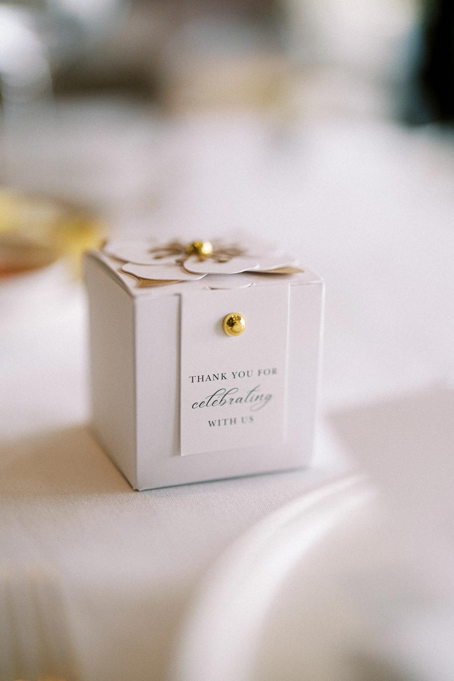 White wedding favor box on a table, adorned with a decorative flower and a gold pin, featuring a Thank you for celebrating