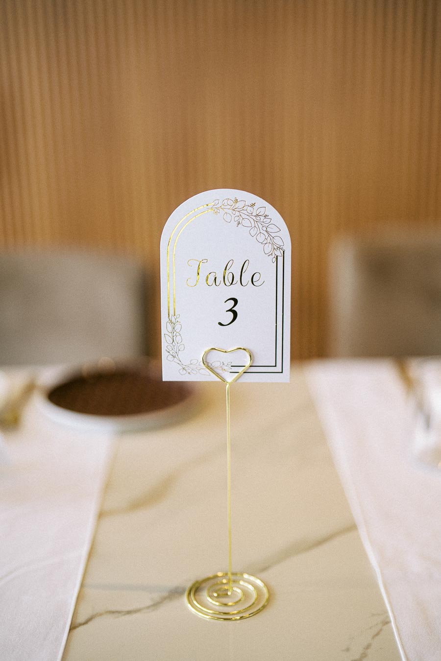 Elegant table number three card on a gold wire stand at a sophisticated event setting with a blurred background.