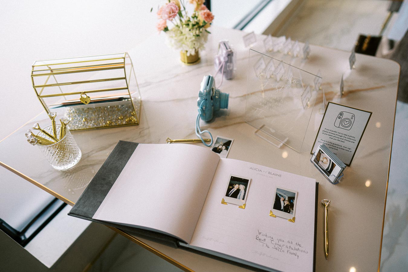Wedding guest book and decor setup on a marble table, featuring a photo album with Polaroid pictures, a gold-pen holder, a
