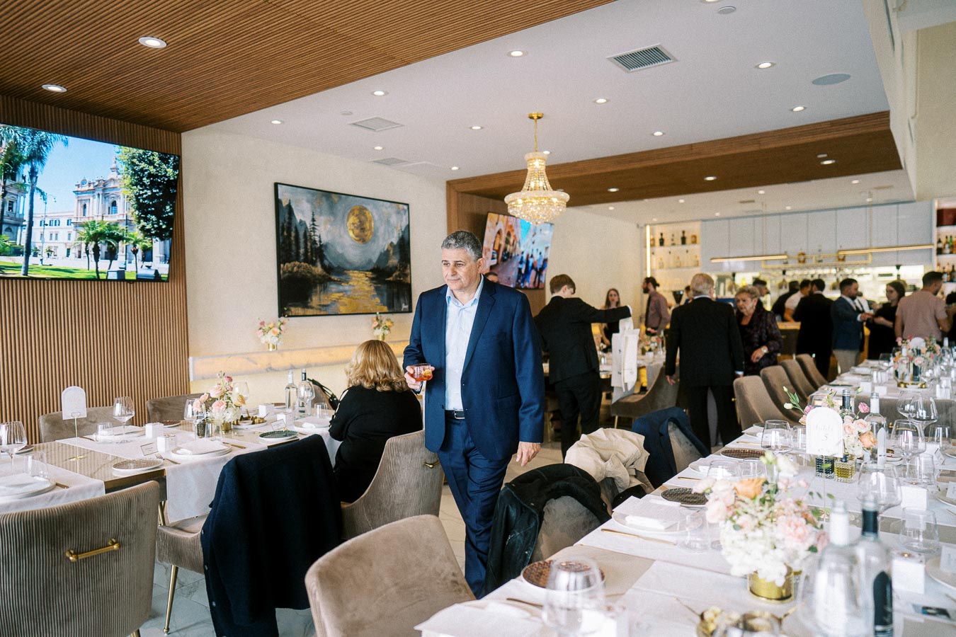Elegant restaurant interior with patrons socializing, featuring beige chairs, white table settings, and floral