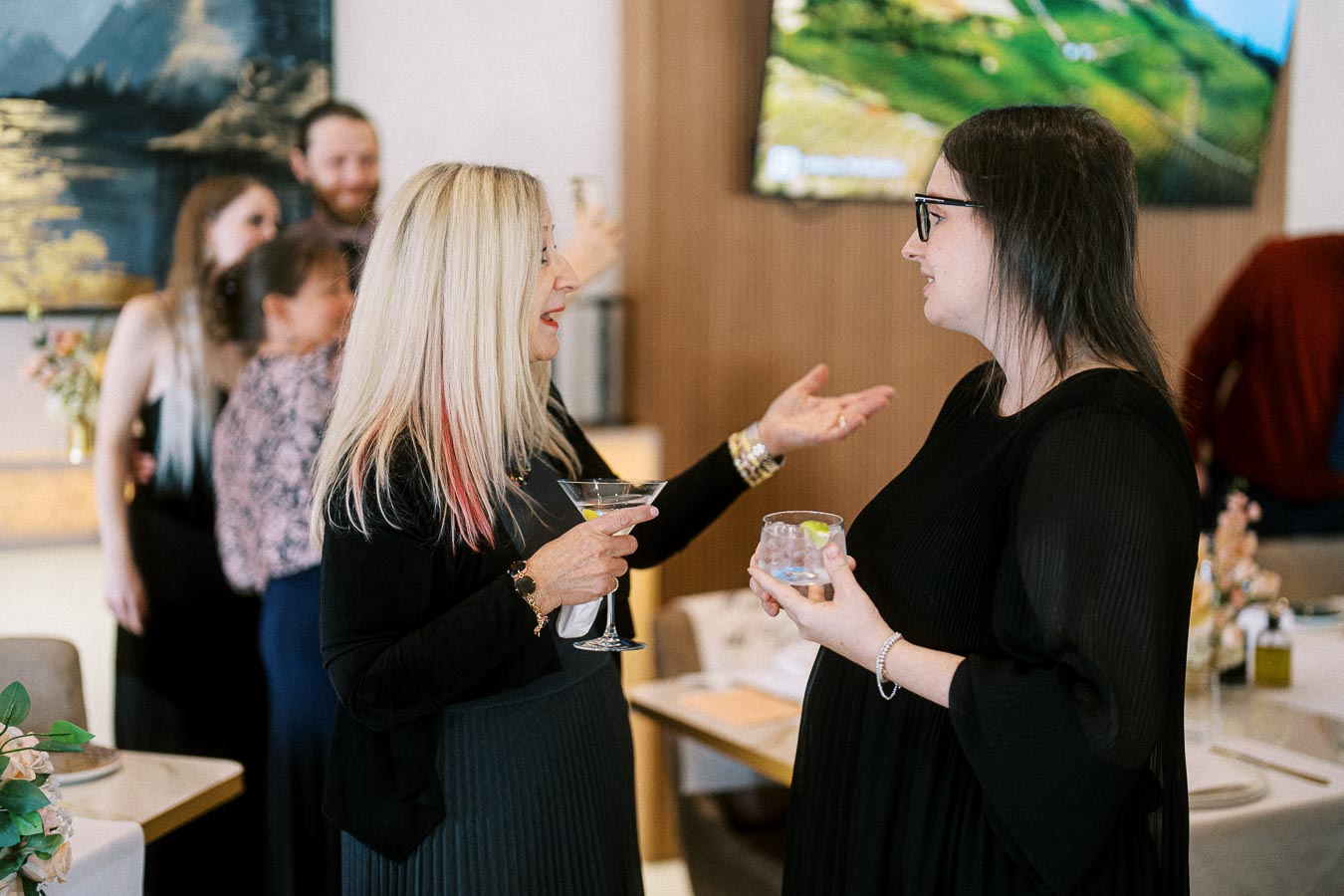 Two women engaged in conversation at a social gathering, each holding a cocktail. In the background, a couple is embracing