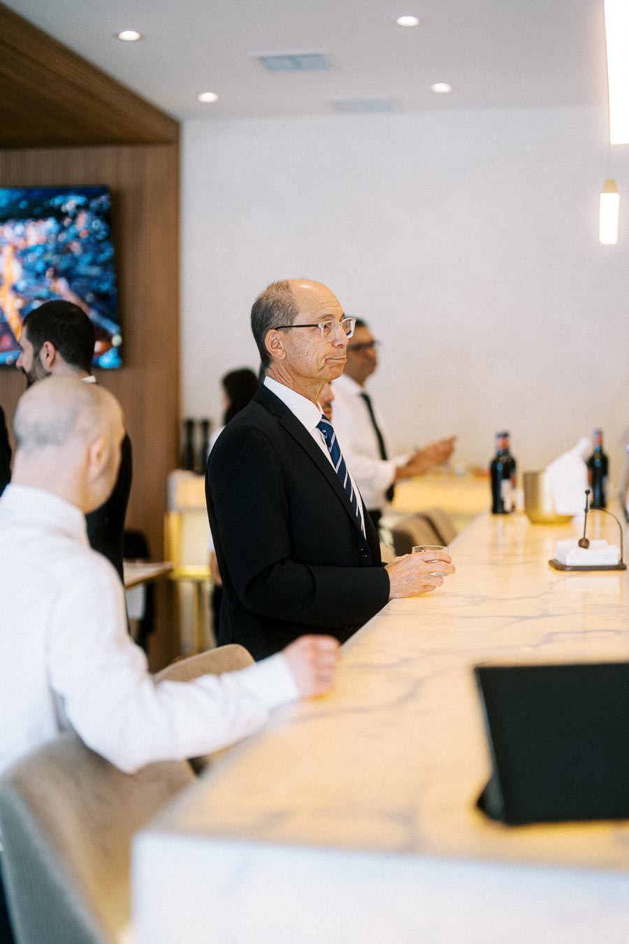A man in a suit stands at a modern bar counter, holding a glass, while other people are visible in the background,