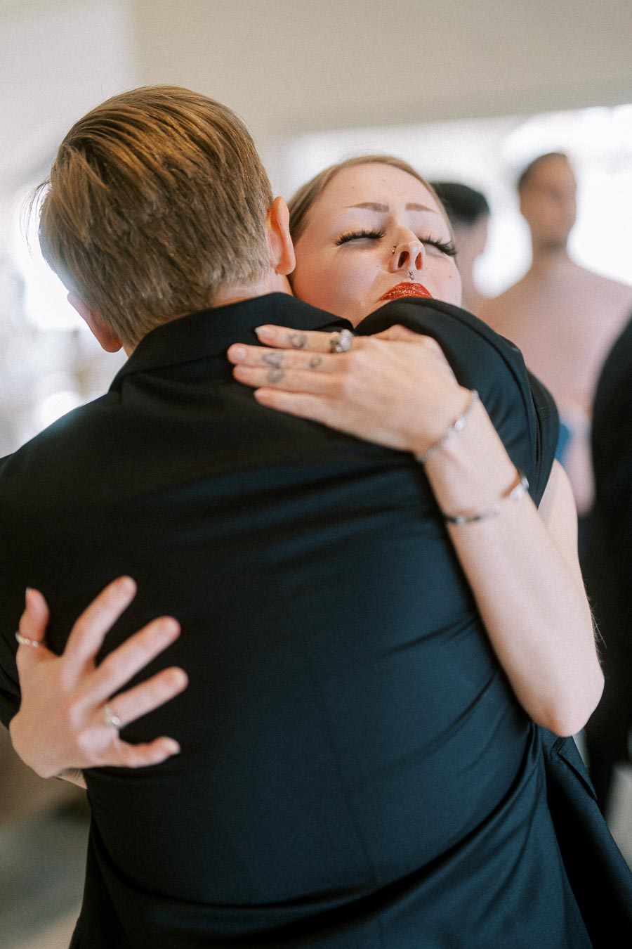 A woman with dramatic makeup and piercings embraces a man in a black suit, capturing an emotional moment.
