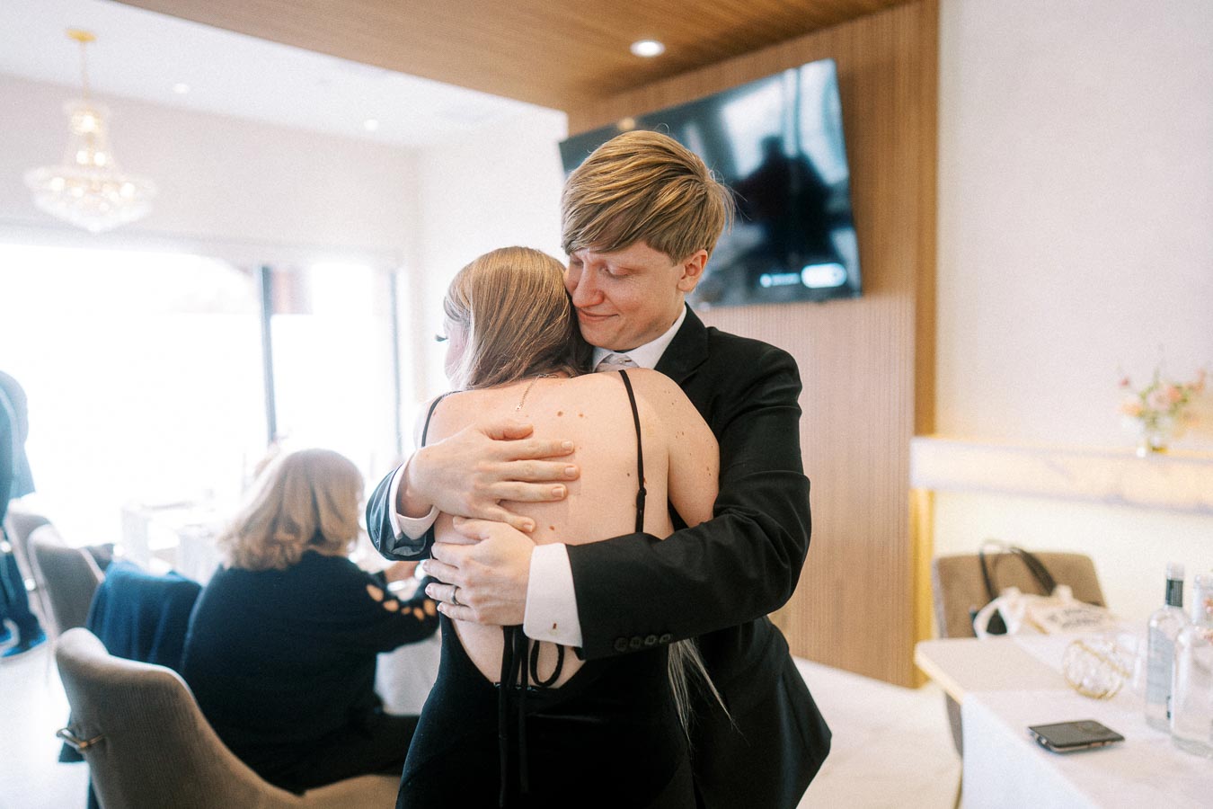 A person in formal attire warmly embraces another individual wearing a black dress in a modern, elegantly decorated room,