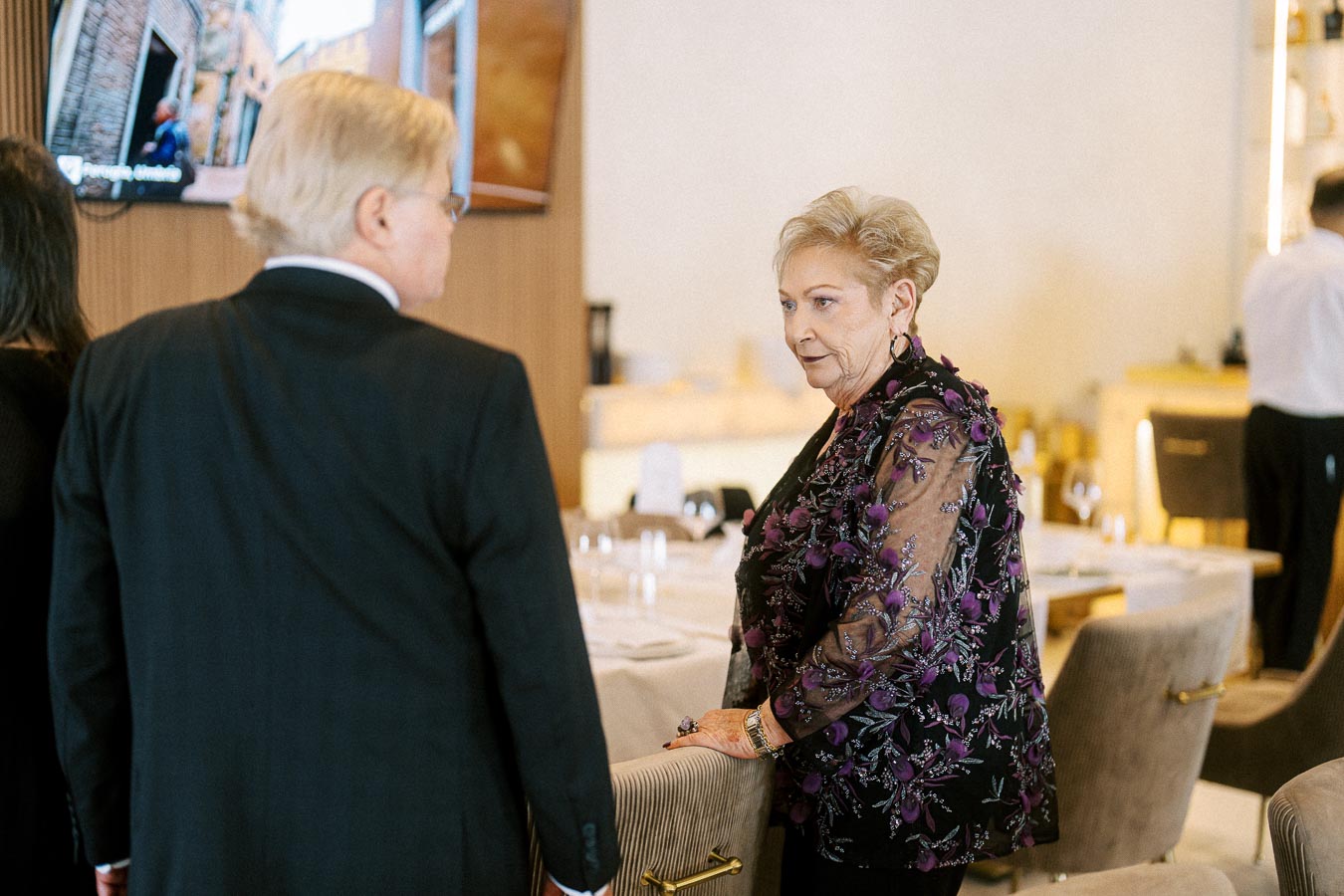 Senior woman in a floral embroidered jacket conversing with a man in a suit at an upscale restaurant setting.