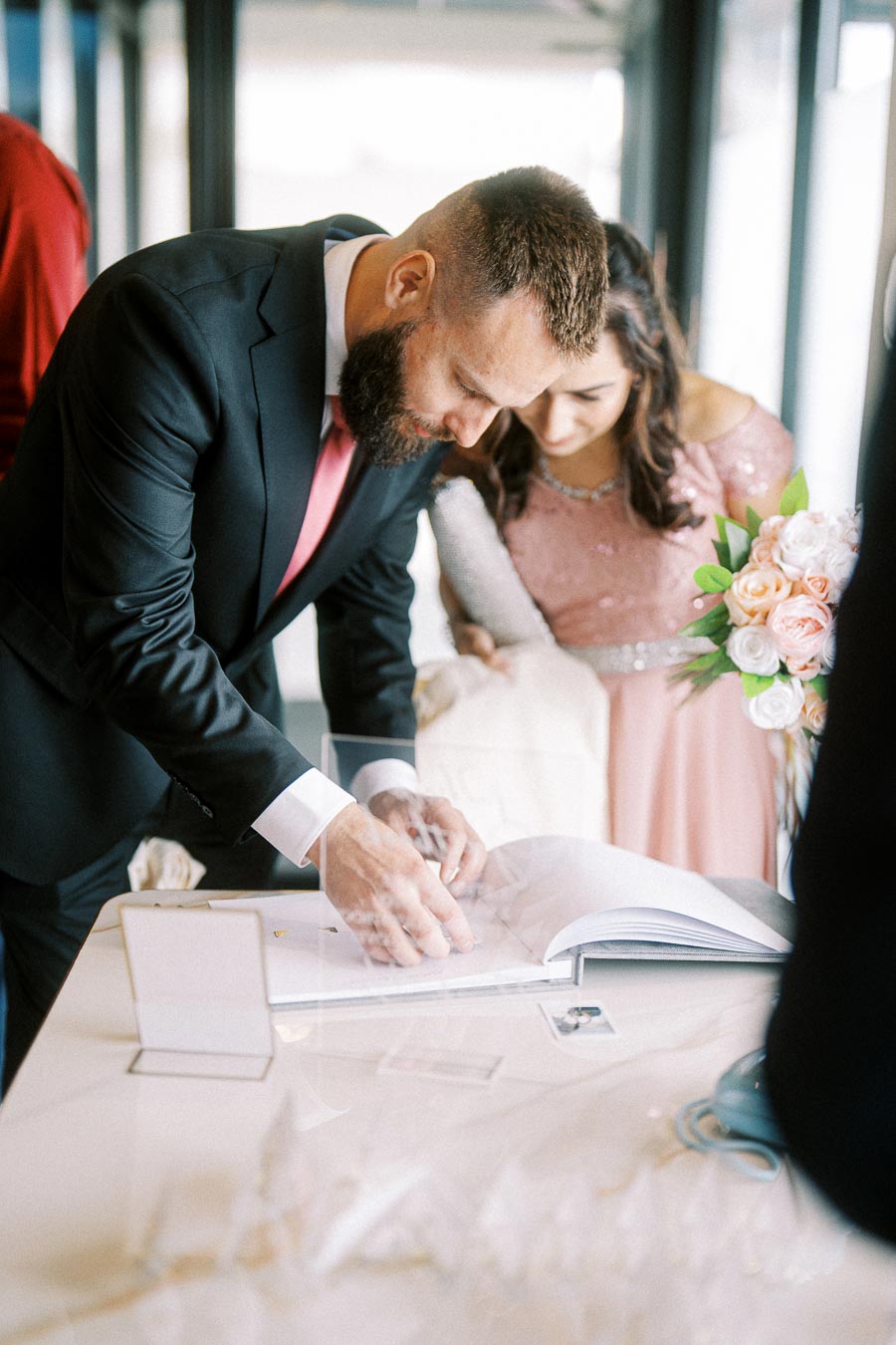 A man in a black suit signing a document at a wedding ceremony table while a woman in a pink dress watches, with a bouquet