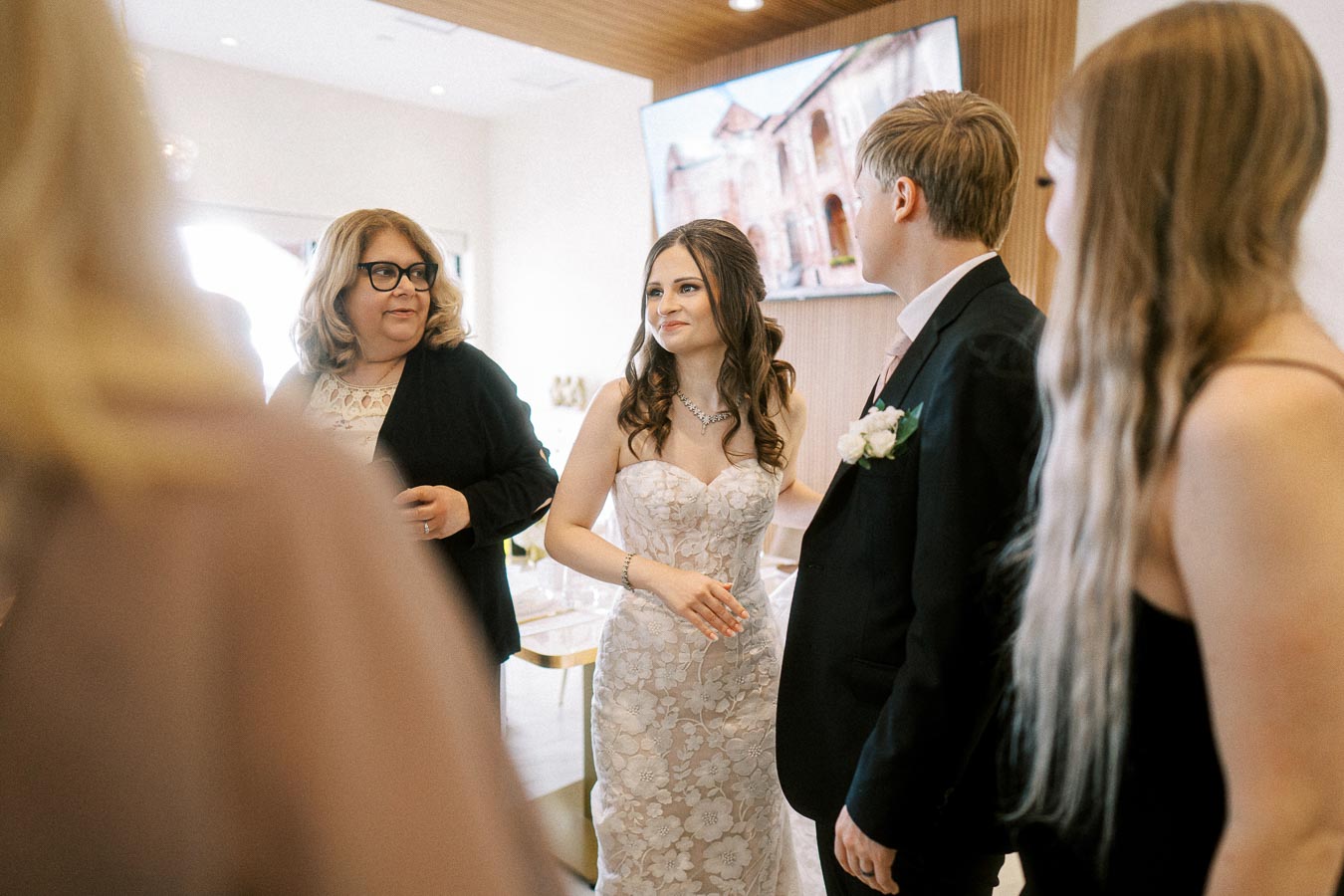 Wedding scene with a bride in a floral lace dress standing with guests, including a woman in glasses and a man in a suit