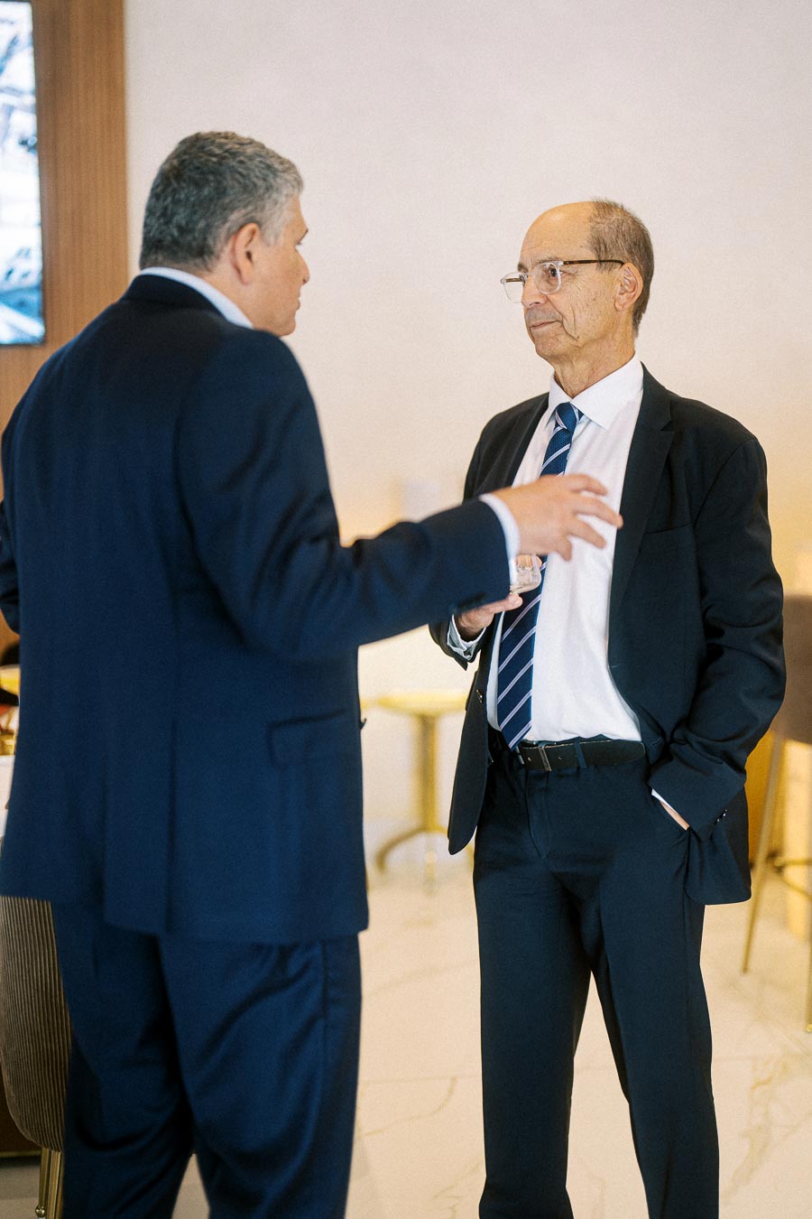 Two businessmen in suits engage in a conversation in a well-lit office setting, with one gesturing while the other listens