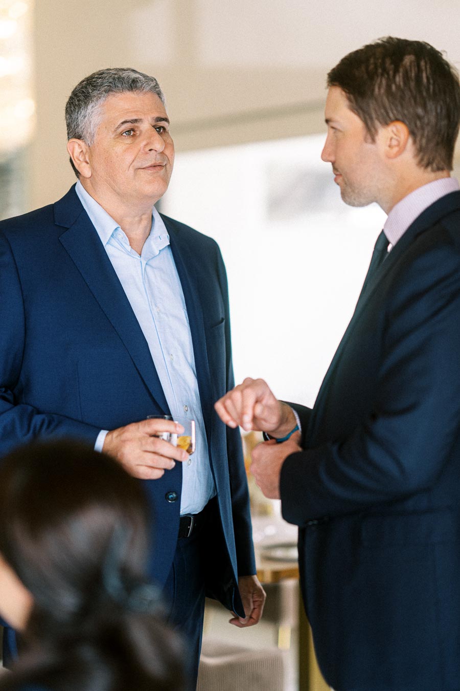 Two men in formal suits engaged in conversation at a professional event. One holds a drink, adding a casual element to the