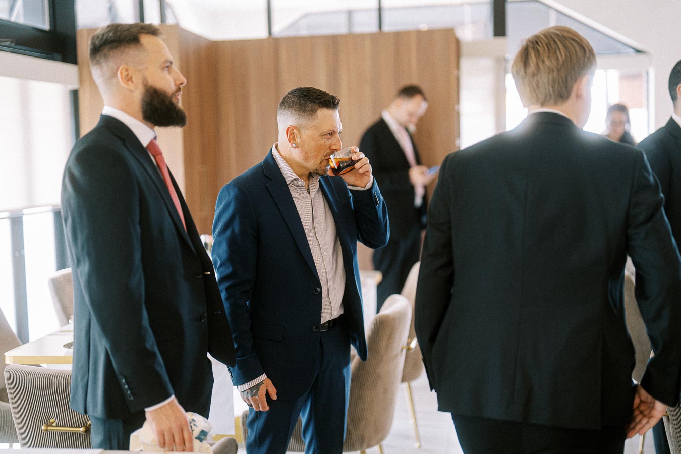 A group of business professionals in formal suits interacting at a networking event, with one man sipping a drink.