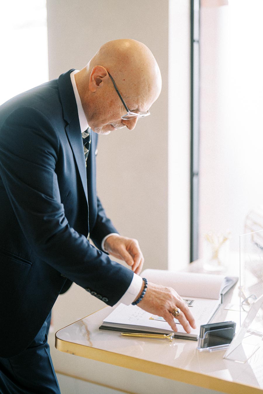 Bald man in a suit writing in a notebook at an office desk with a pen.